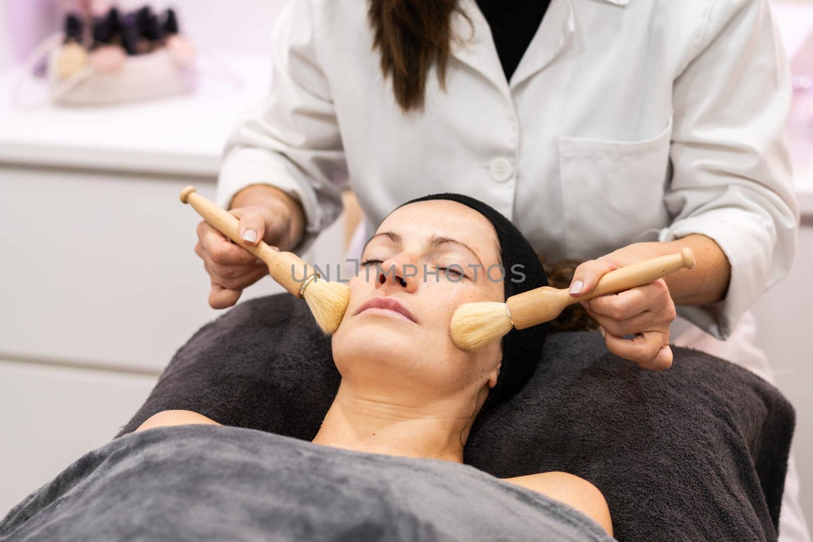 Crop unrecognizable cosmetician in uniform massaging face of female customer with brushes during facial treatment at beauty salon