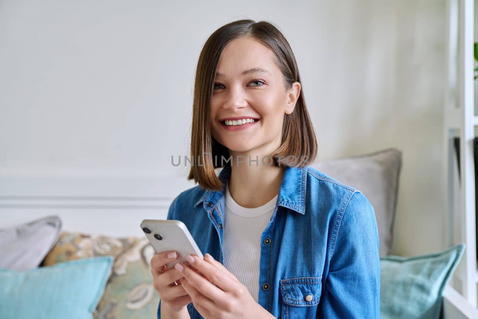 Young smiling woman using smartphone sitting on couch at home. Female looking at camera texting reading looking mobile applications technology internet online services for work study leisure lifestyle