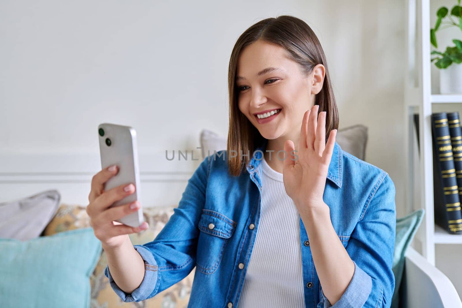 Young woman sitting with smartphone on couch at home having video call. Smiling female chatting with friends colleagues on social networks, student talking in online lesson freelancer working remotely