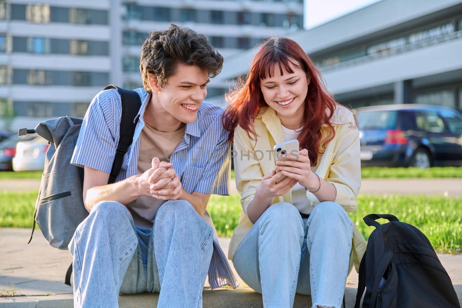 Friends young guy and girl university college students talking together, looking at smartphone outdoor, city modern buildings background. Lifestyle, communication, youth 19-20 years old, urban style