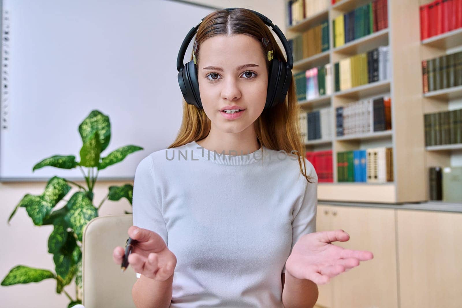 Webcam view of teenage girl, high school student, in headphones, talking to camera. Female teenager studying remotely, video conference chat, online test exam lesson. Technology education adolescence