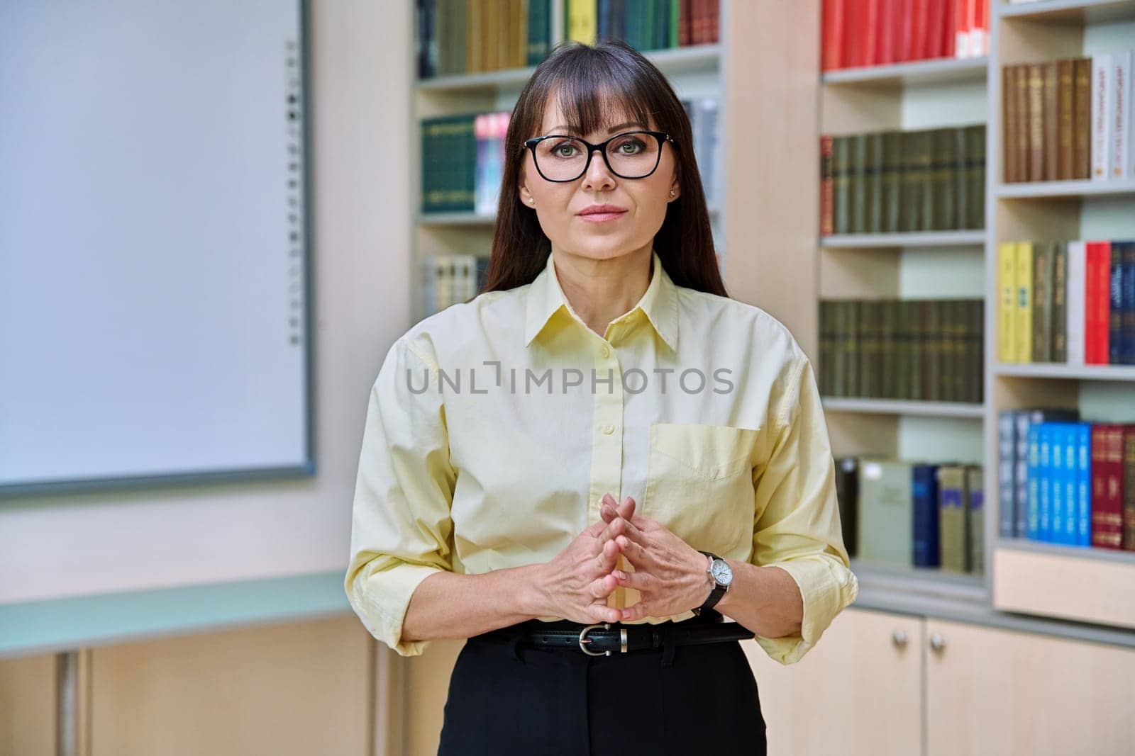 Portrait of confident mature business woman in library. Middle-aged elegant female with folded hands, manager teacher mentor psychologist counselor librarian advisor background of shelves with books