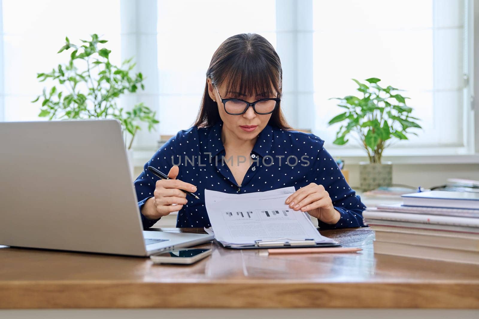 Middle-aged serious woman working with laptop computer with business papers, sitting at desk in home office interior. Work, remote business, freelance, technology, 40s people concept