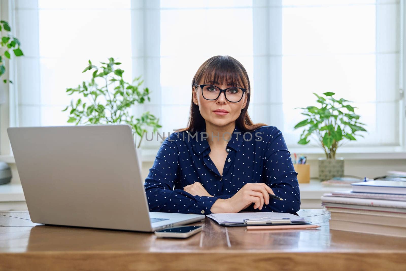 Middle-aged serious woman working with laptop computer business papers, sitting at desk in home office interior, looking at camera. Work, remote business, freelance, technology, 40s people concept