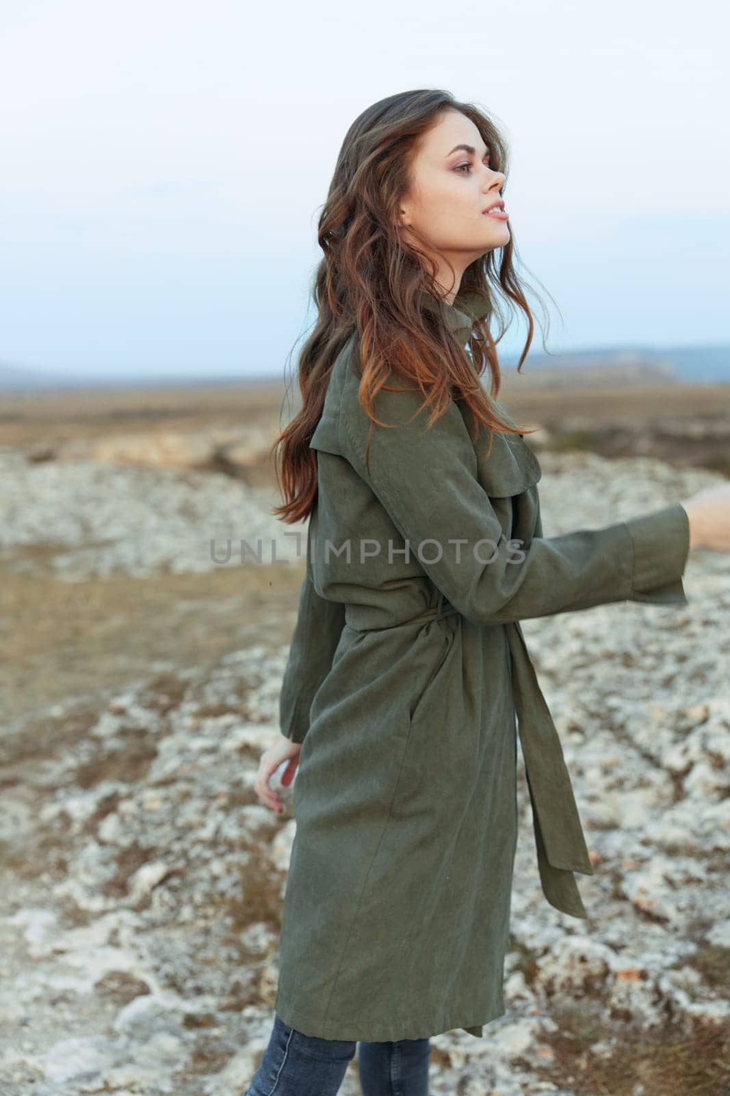 Woman in olive green trench coat standing on rocky hill overlooking ocean on sunny day