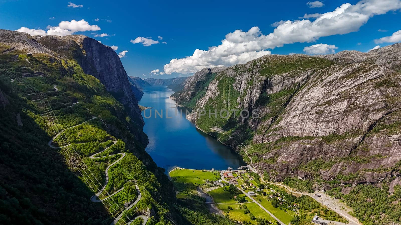 An aerial view of a winding road snaking through the dramatic, green cliffs of a Norwegian fjord on a bright, sunny day. Kjerag, Lysebotn, Lysefjorden, Norway