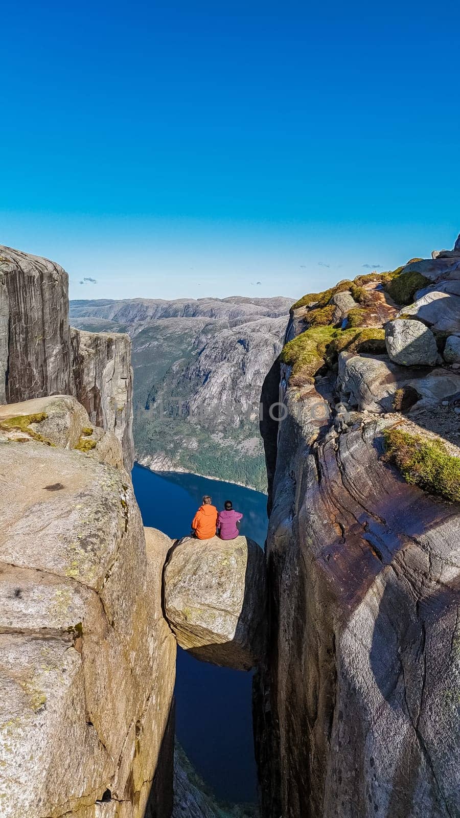 Two individuals sit on a narrow rock ledge overlooking a deep valley in Norway. The majestic Kjeragbolten cliff provides a breathtaking backdrop for this daring feat. a couple at Kjeragbolten