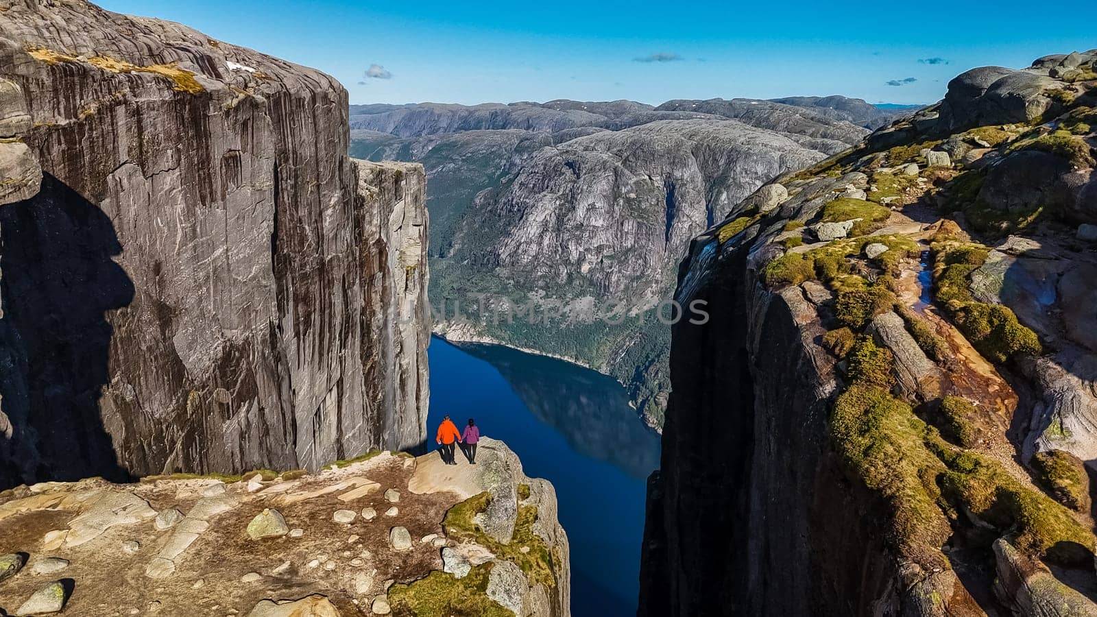 Two hikers stand on the edge of Kjeragbolten, Norway, a famous cliff in Norway, with a breathtaking view of the surrounding fjord. couple of men and women visit Kjeragbolten, Norway