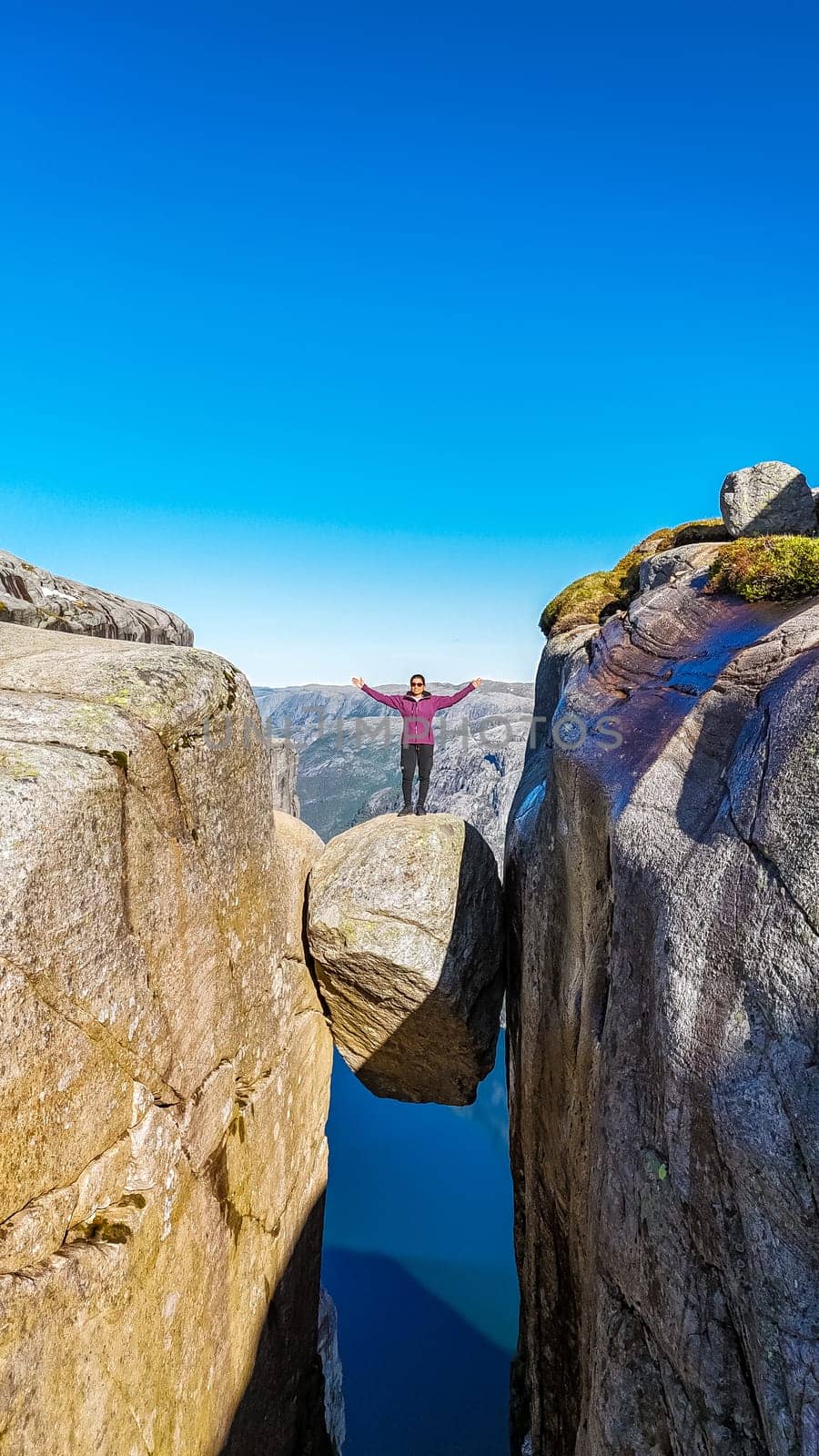 A person stands with arms outstretched on a rock formation at Kjeragbolten, Norway, on a clear day. Asian women with hands up at Kjeragbolten, Norway