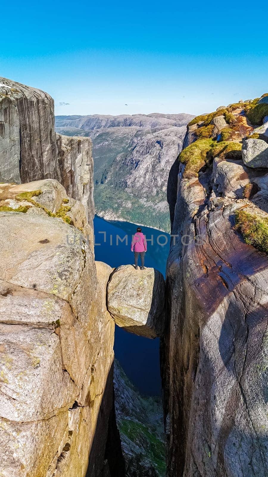 A lone hiker stands on the edge of Kjeragbolten, a cliff in Norway, looking out over the vast Norwegian landscape.