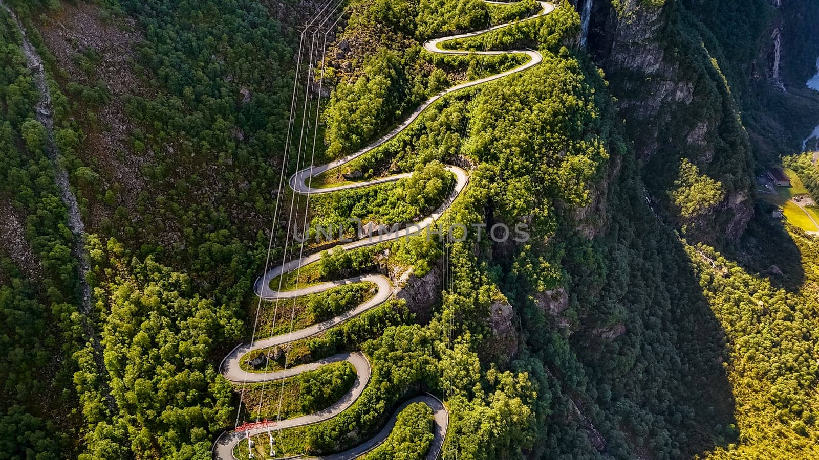 An aerial view of a winding road through the mountains of Norway. The road is surrounded by lush green trees and slopes down into a valley below. Lysebotn, Lysefjord, Norway