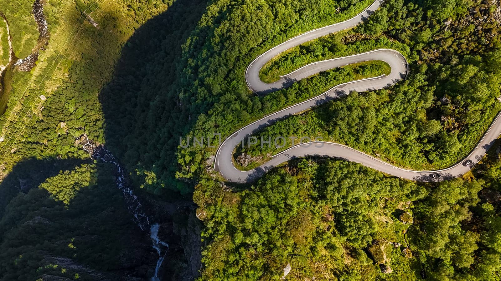 An aerial view of a winding road snaking through a dense forest in Norway. The roads curves and the vibrant green foliage create a stunning visual.