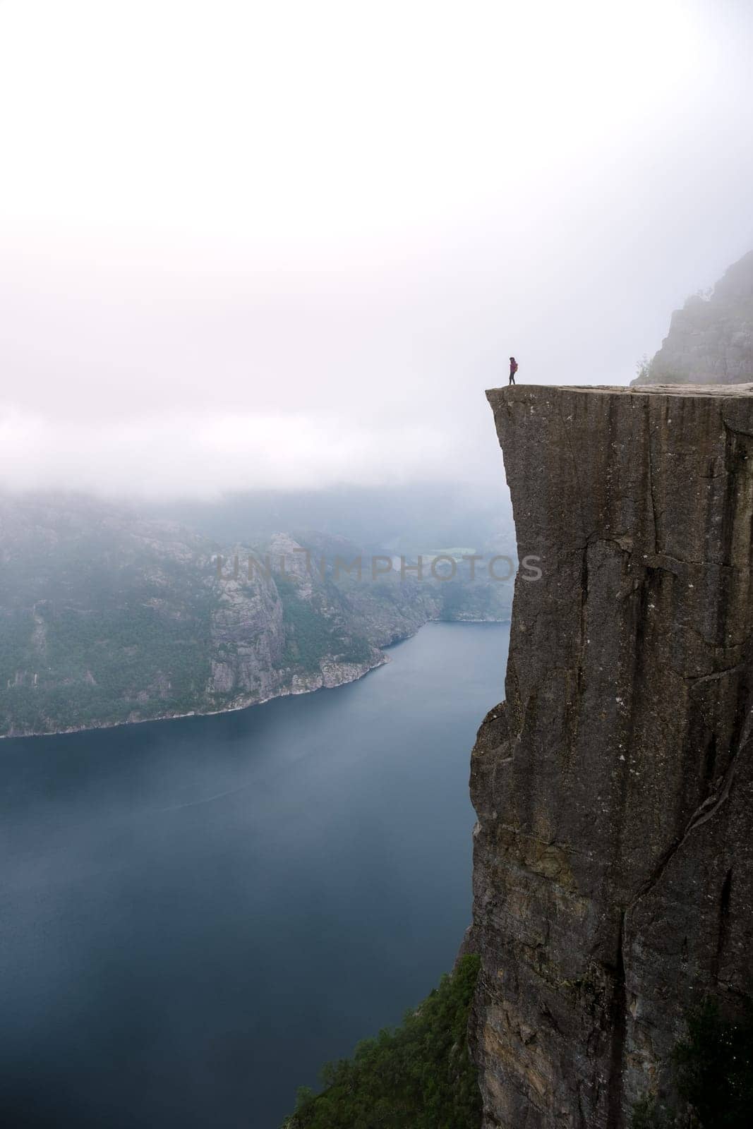 A lone individual stands on the edge of Preikestolen, a cliff in Norway overlooking a fjord. The sky is misty and the landscape is dramatic. Preikestolen, Norway