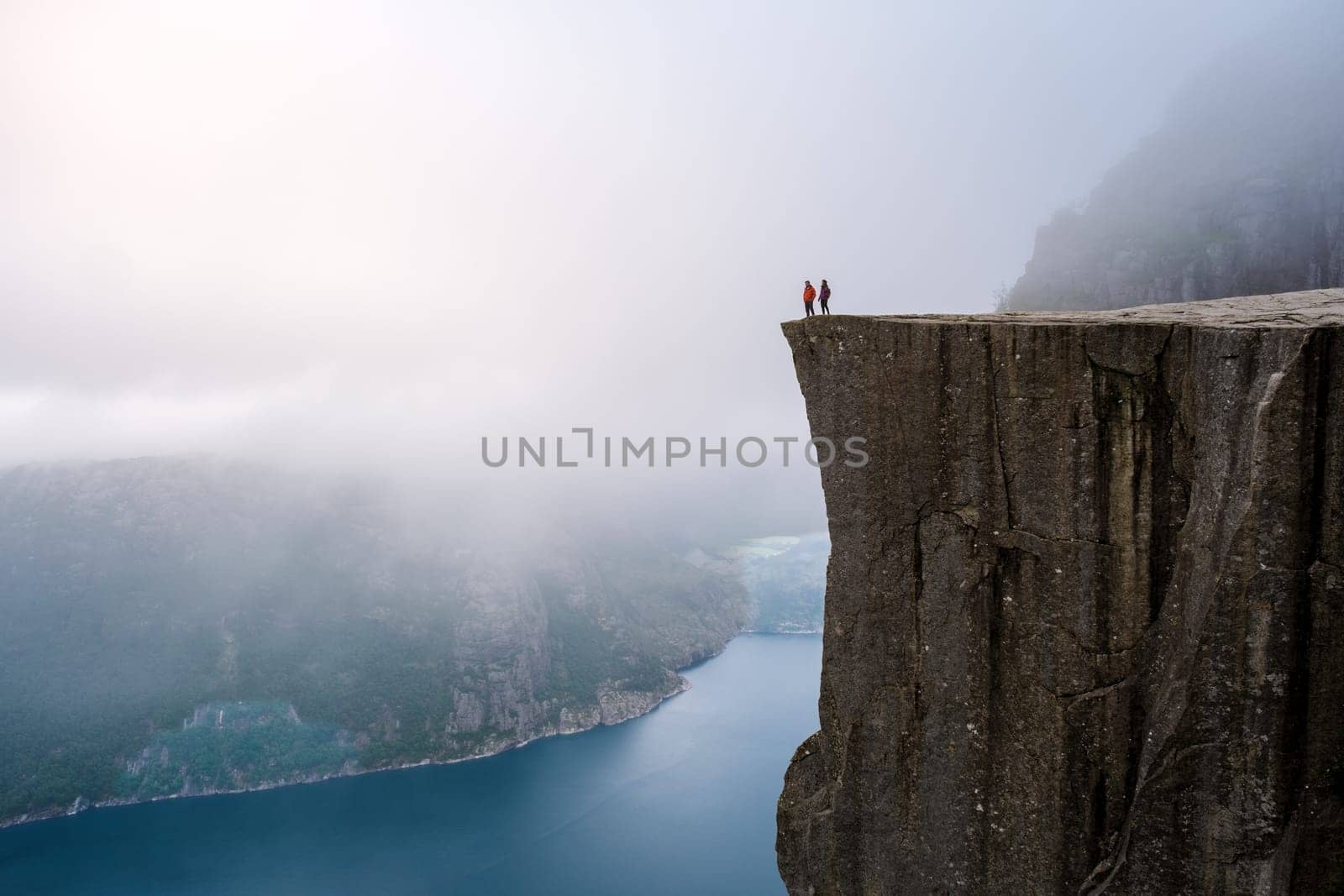 A view from the top of the Pulpit Rock in Norway, with two figures standing on the edge, looking out over the misty fjord. Preikestolen, Norway