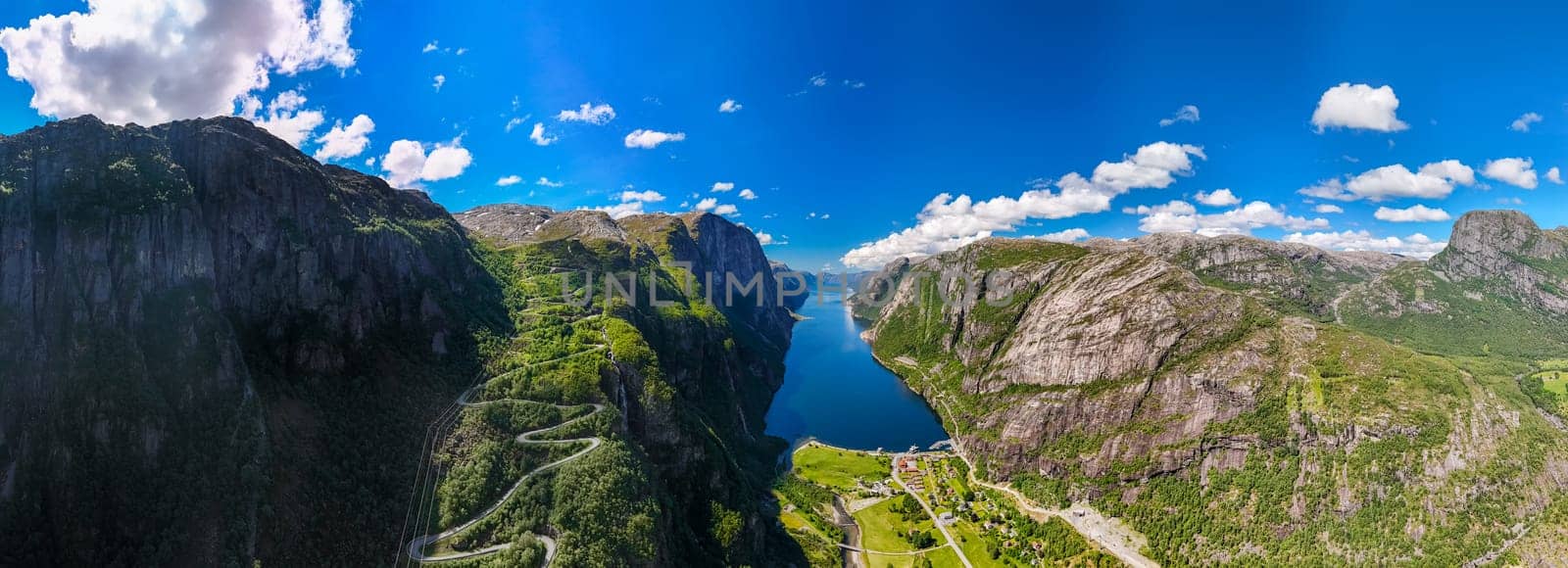 An aerial view of a winding road snaking through the towering mountains and lush greenery of a Norwegian fjord, with a clear blue sky and fluffy white clouds. Lysebotn, Lysefjorden, Norway