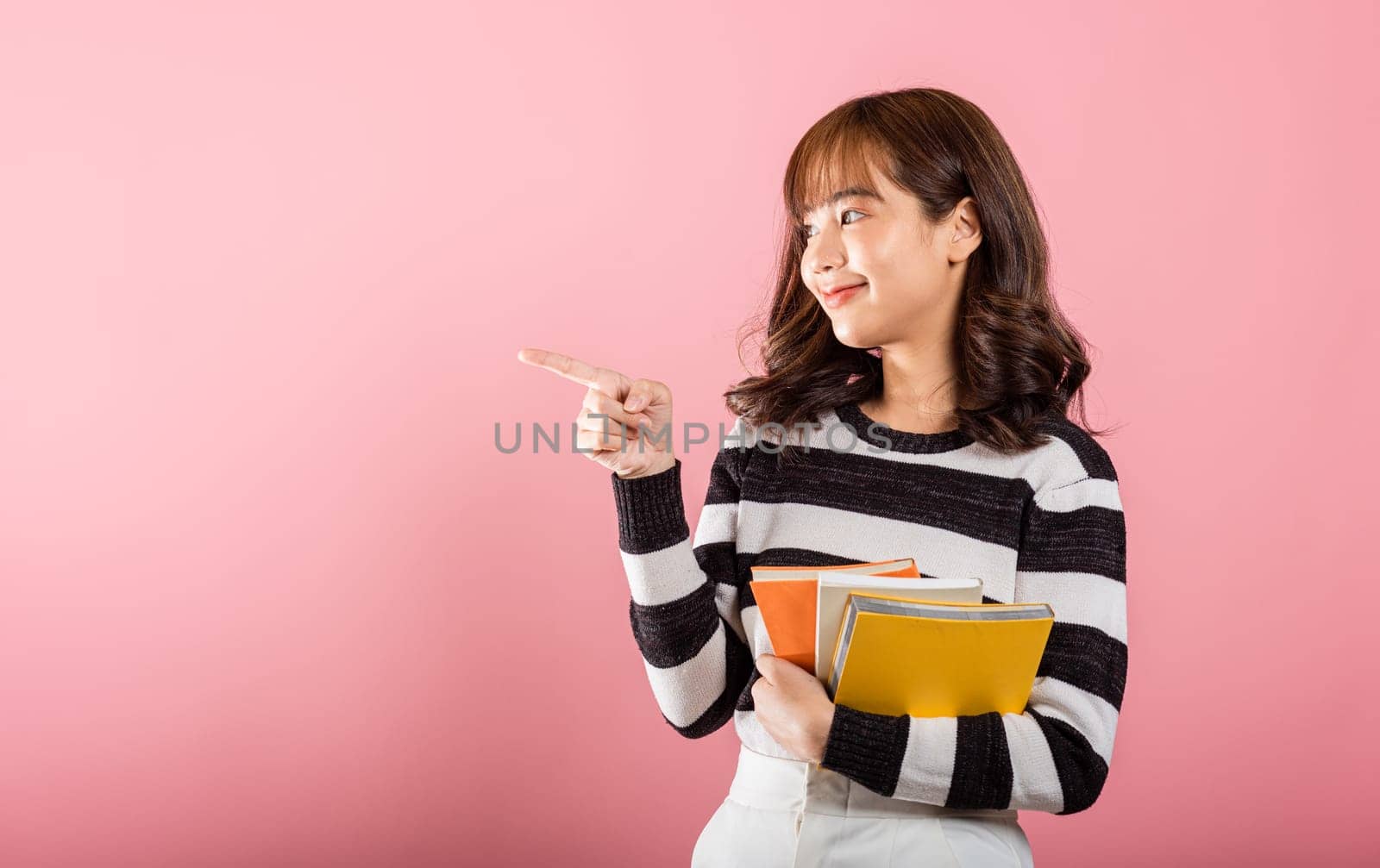 Portrait of happy Asian beautiful young woman confident smiling holding orange book open pointing finger to side copy space, studio shot isolated on pink background, education concept