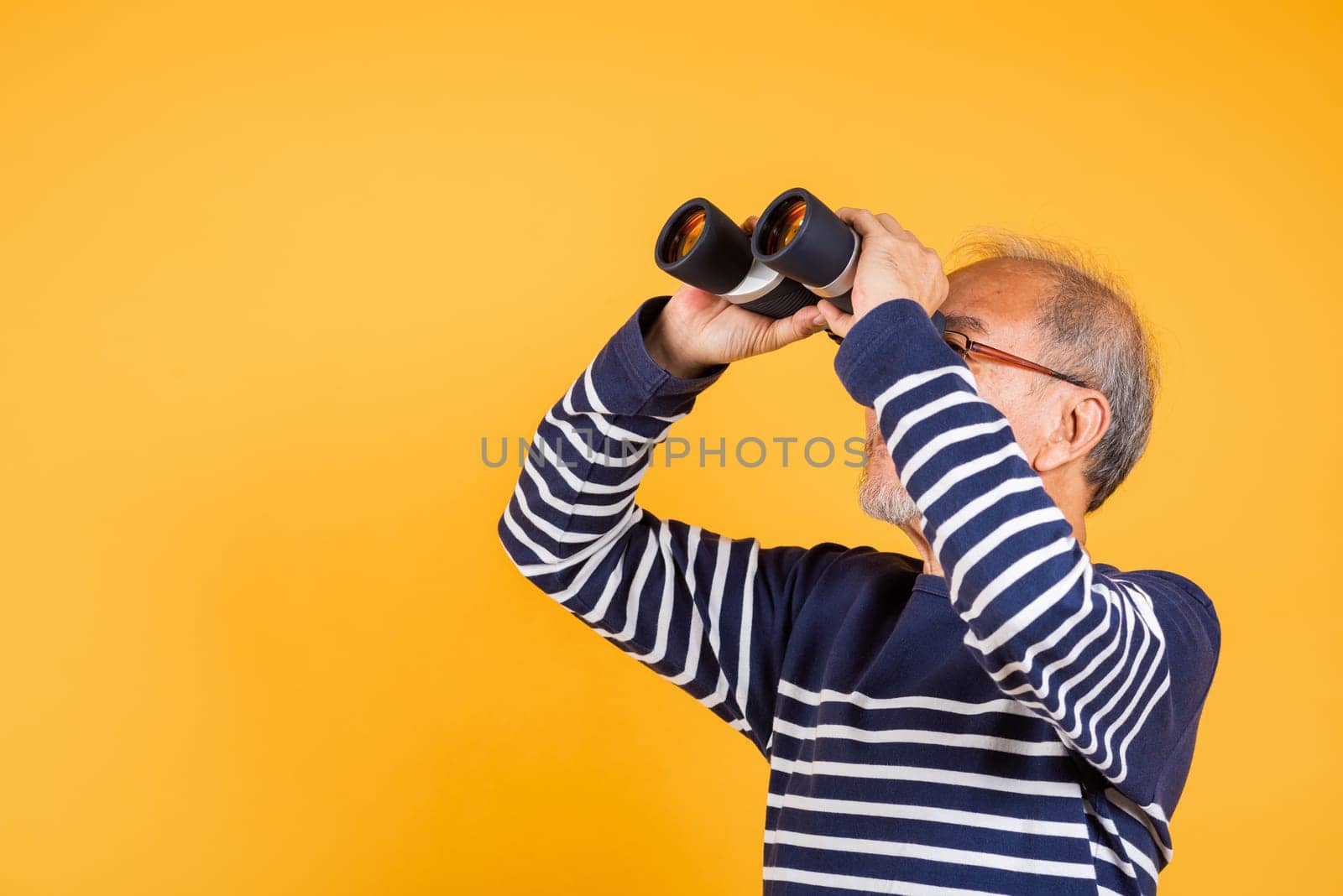 Portrait Asian smiling old man looking camera through binoculars studio shot isolated yellow background, senior man pensioner feeling surprised using binocular