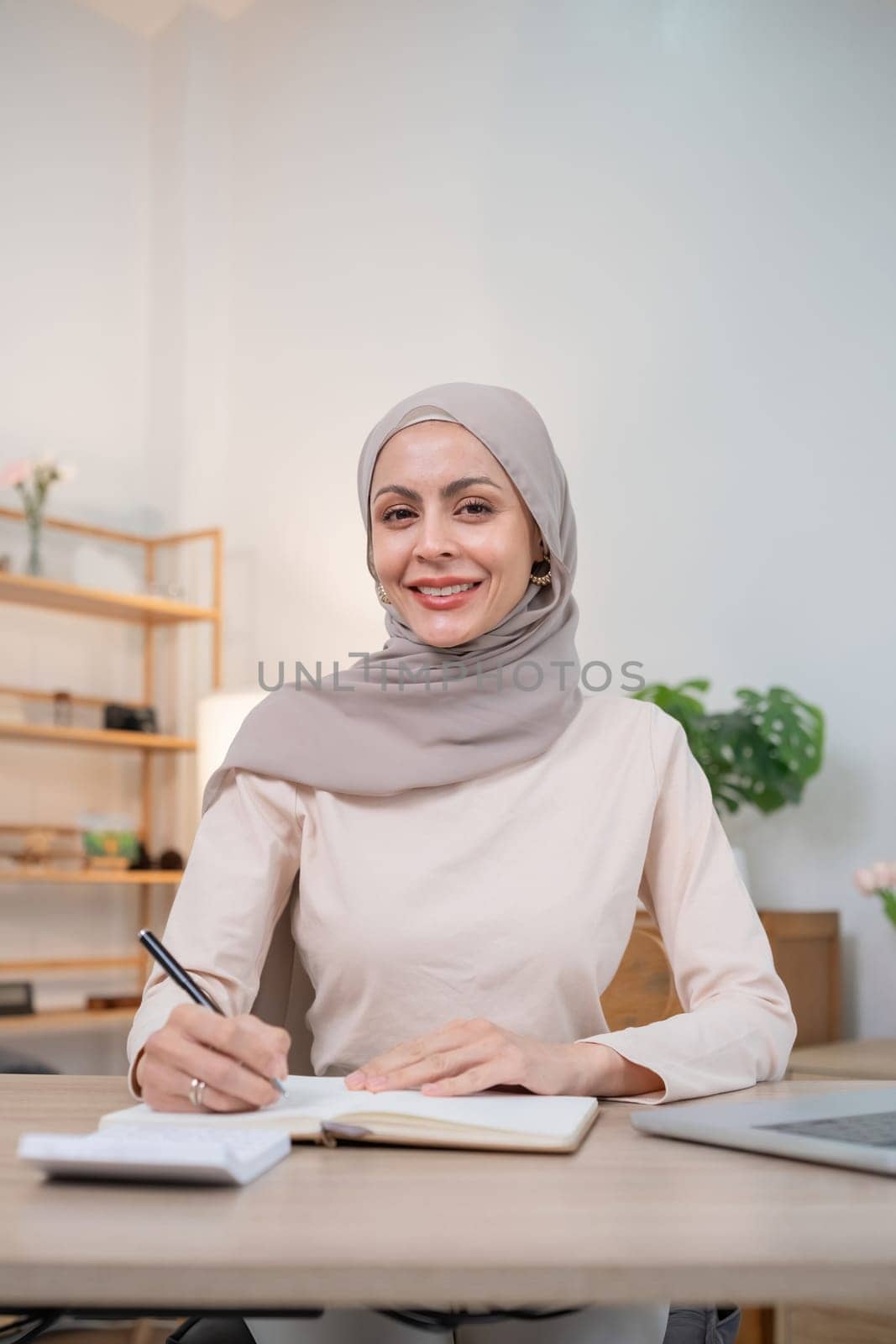 A confident Muslim woman in a hijab working at a desk, smiling and writing in a notebook, representing professionalism and empowerment in a modern office environment.