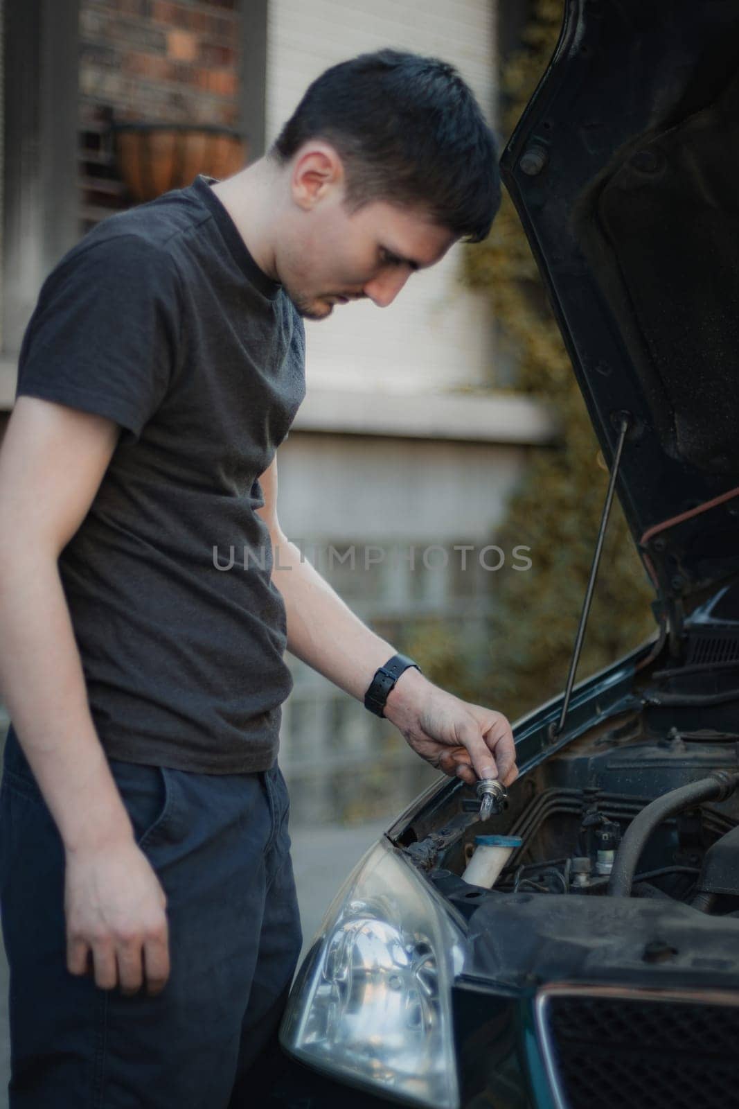 One young Caucasian guy holding in his hand a burnt-out lamp from the right front headlight of a car, standing half-turn on a city street on a summer day in the evening, close-up side view. Concept for replacing light bulbs, car repair.