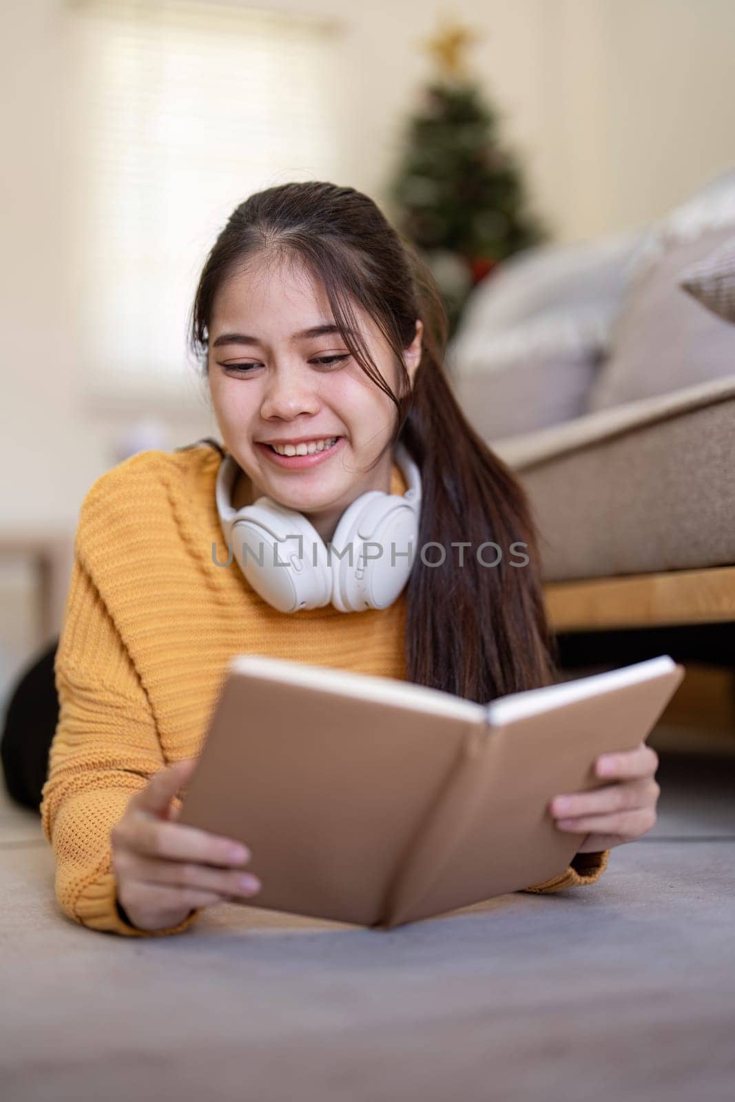 A young woman reading a book at home, lying on the floor with headphones around her neck, enjoying a cozy and comfortable indoor setting.