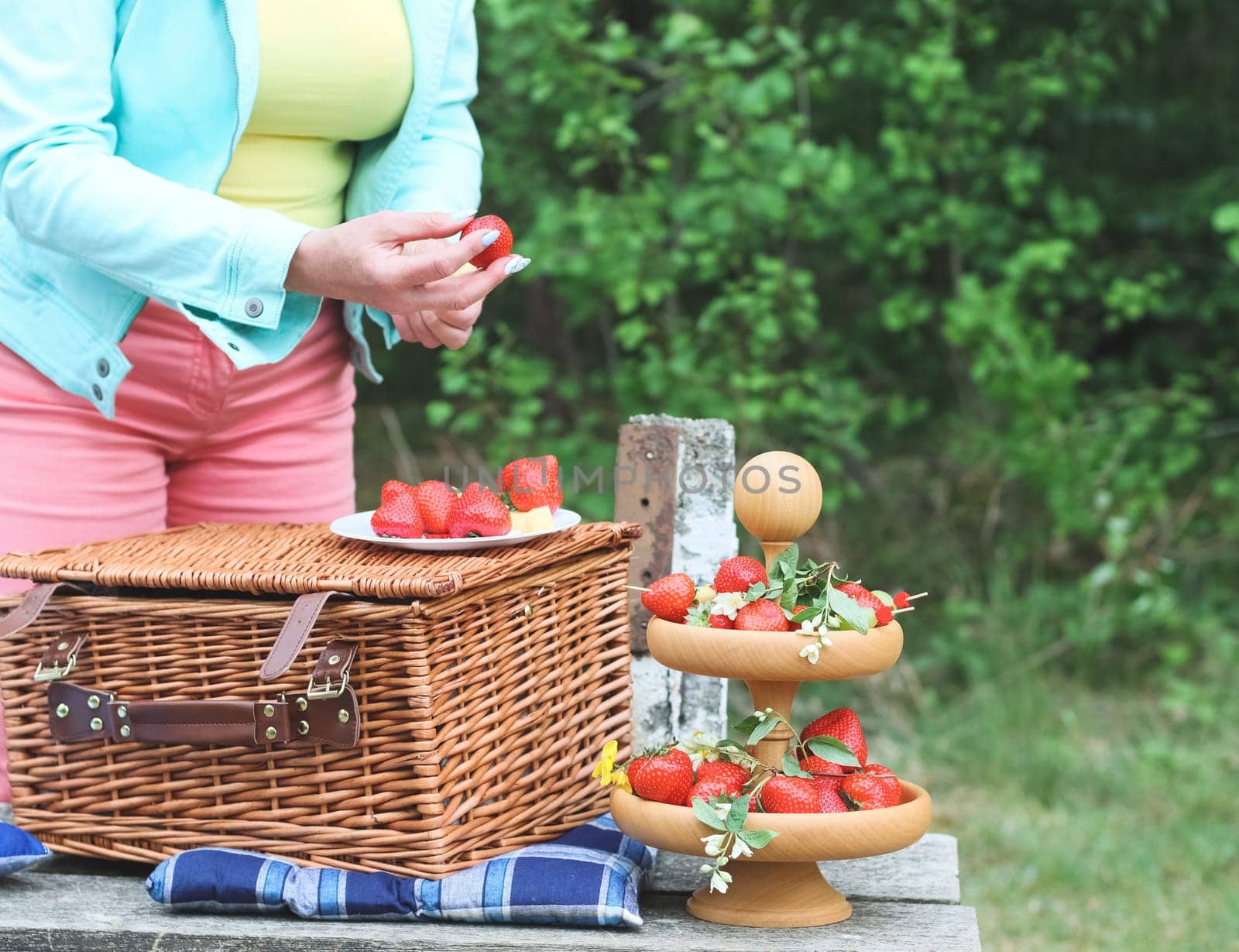 The hands of a middle-aged caucasian woman prepare fruit skewers of strawberries, grapes and cheese, skewering them on wooden skewers, standing near a bench with a wicker basket in the forest, close up side view. Concept picnic outdoor recreation.