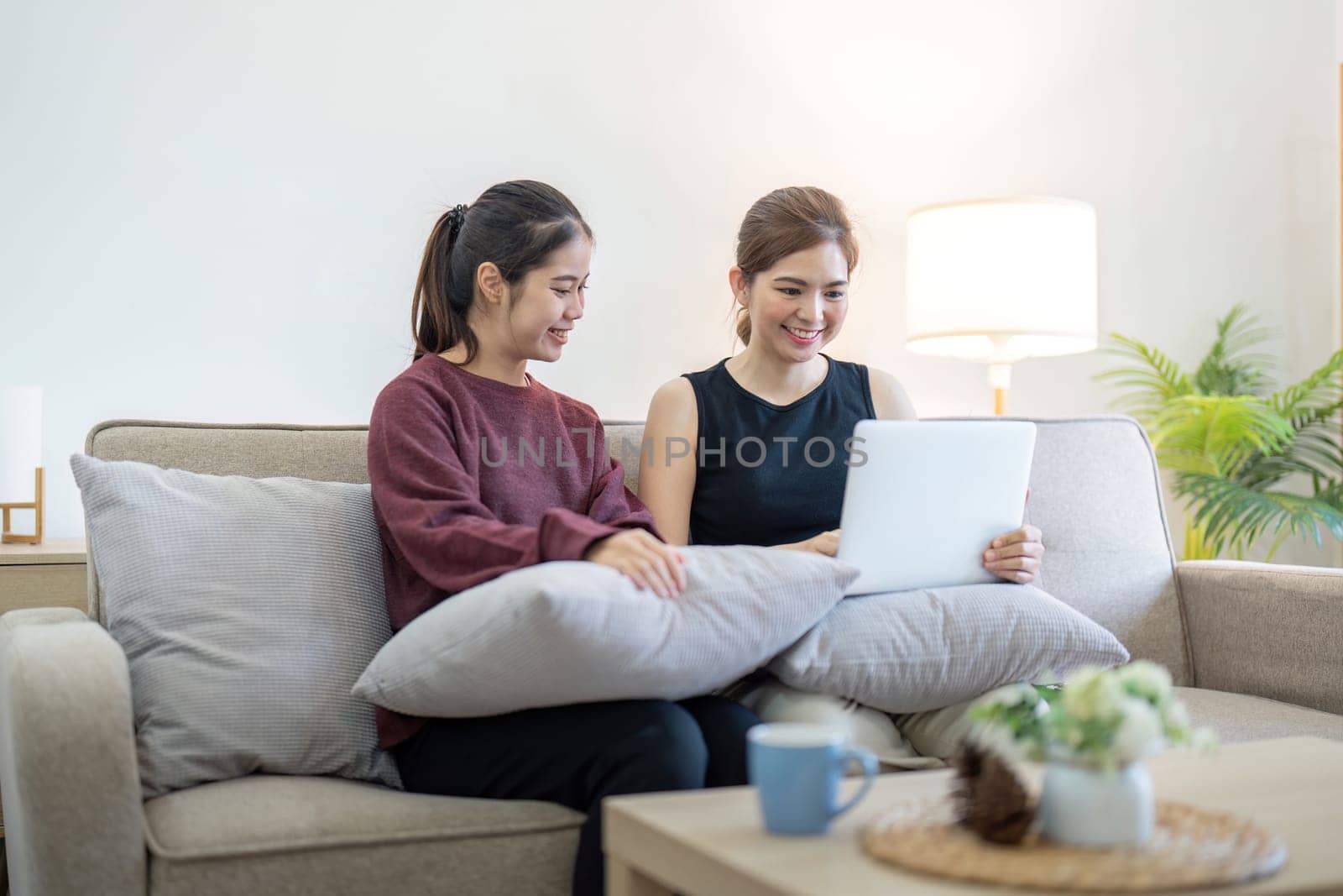 Lesbian couple sitting on a couch, using a laptop, and smiling. Modern living room setting with casual clothing, representing LGBTQ community and love.