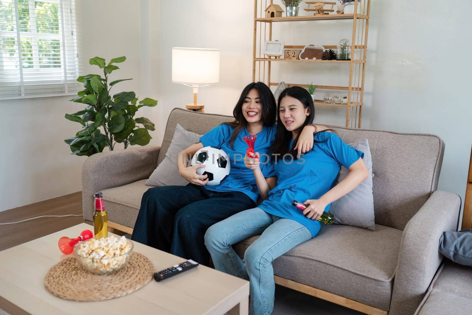 Couple woman friend having fun watching football match on TV, drinking beer and cheering football fans watching game at home celebrating after their team scoring a goal.