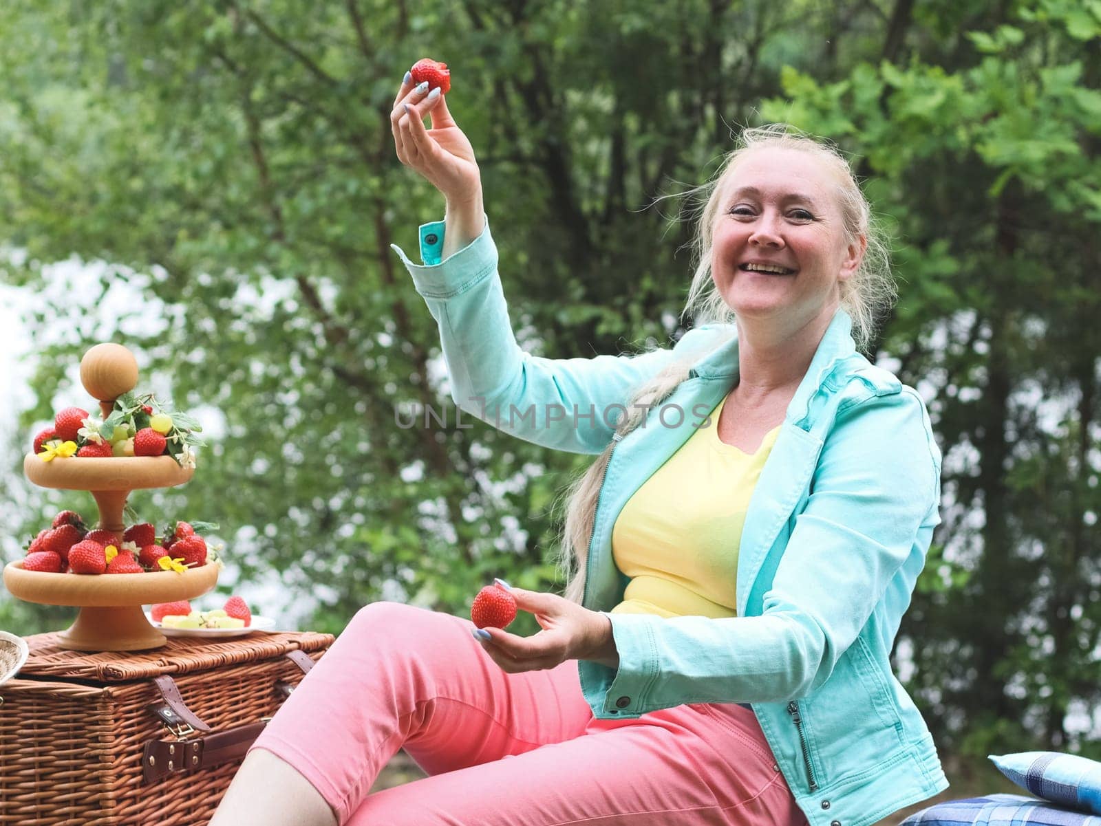 Portrait of a beautiful Caucasian cheerful middle-aged blonde woman, fooling around and having fun, eating ripe strawberries, sitting in a park on a bench on a spring day, close-up side view. Outdoor picnic concept, healthy eating and lifestyle.