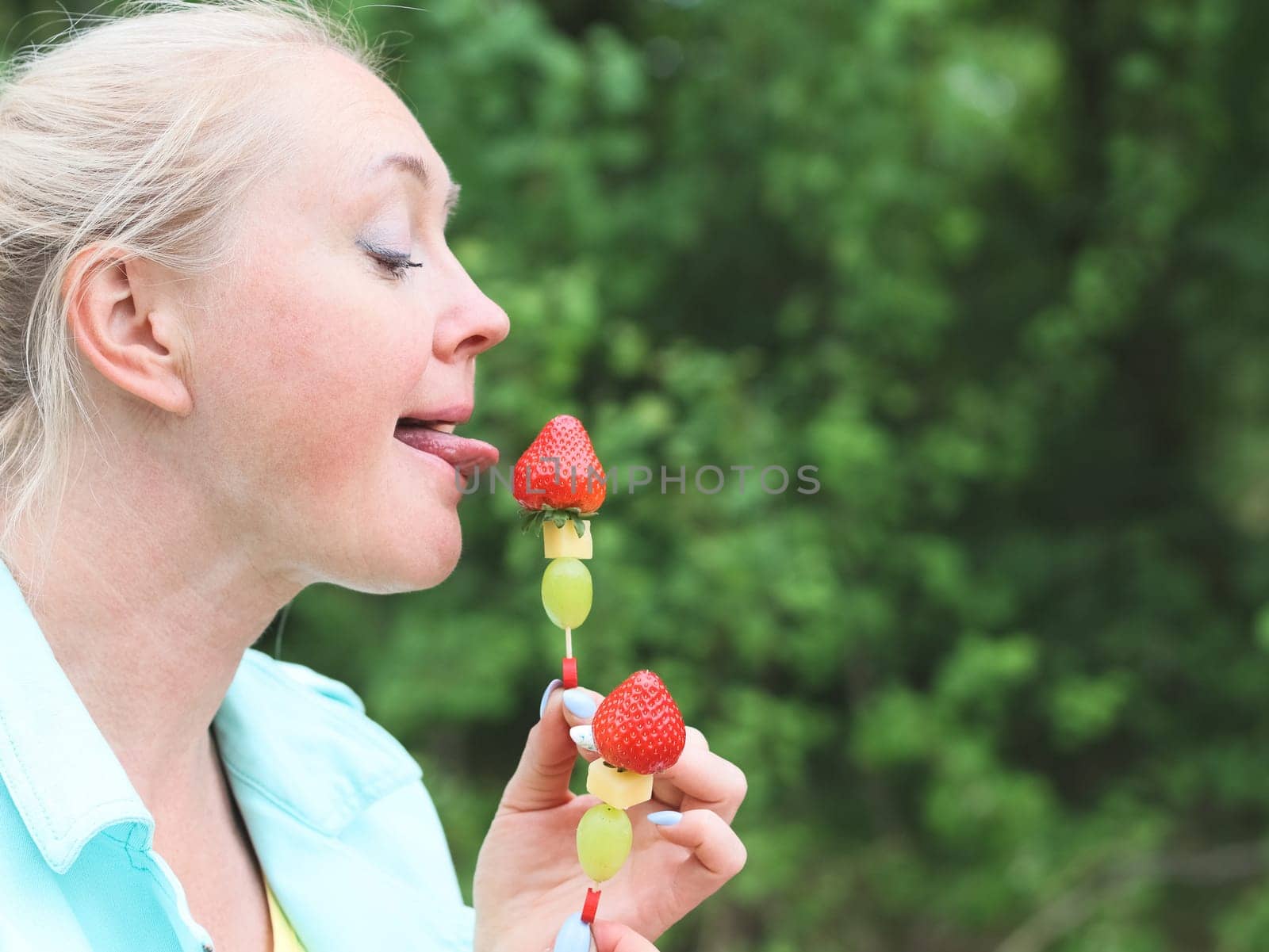 One Caucasian young beautiful blonde woman with delicate manicure in a blue jacket and yellow T-shirt is holding licking two fruit barbecue skewer of strawberries, cheese and wongrad in the park on a spring day, bottom close-up view with copy space on the right. Spring picnic concept, healthy eating.