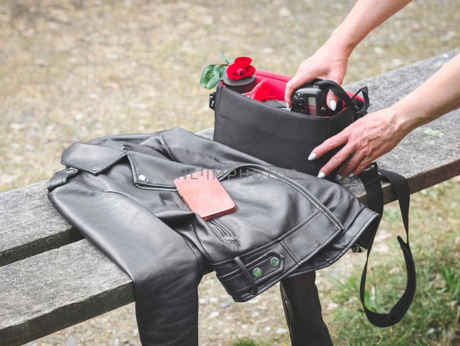 The hands of a young Caucasian woman with a beautiful delicate manicure take out a camera with a lens and a red rose from her purse on a wooden bench with a black leather jacket and a pink smartphone in a public park, close-up side view. Concept using technology, photography, theft.