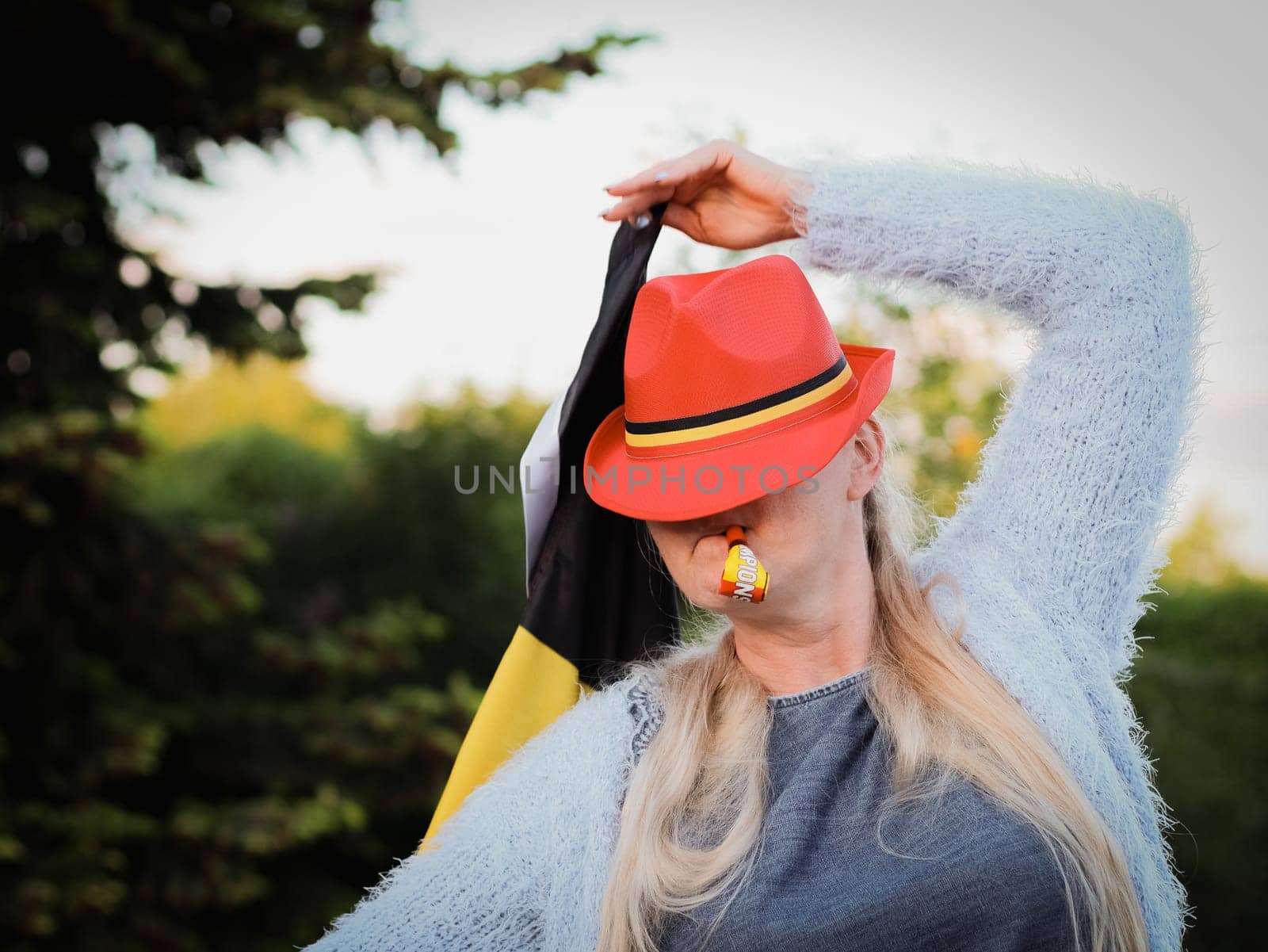 Portrait of a beautiful middle aged blonde caucasian woman with a cap on her head holding a Belgian flag and blowing a whistle celebrating independence day and winning a football match in the backyard of her house,side view closeup.Belgium day concept,flags,symbols,celebration,football,moms , fans.