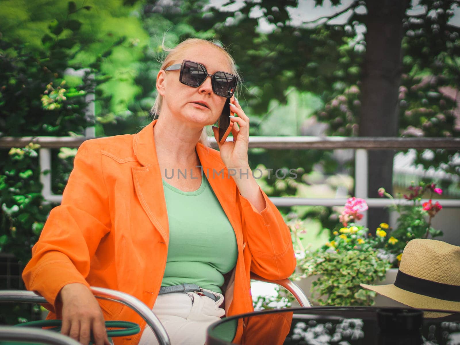 A young beautiful stylish Caucasian blonde woman in sunglasses, a bright orange jacket talks using a mobile phone, sits with her legs stretched out on a chair in a street cafe on a summer day, close-up side view. Technology use concept.