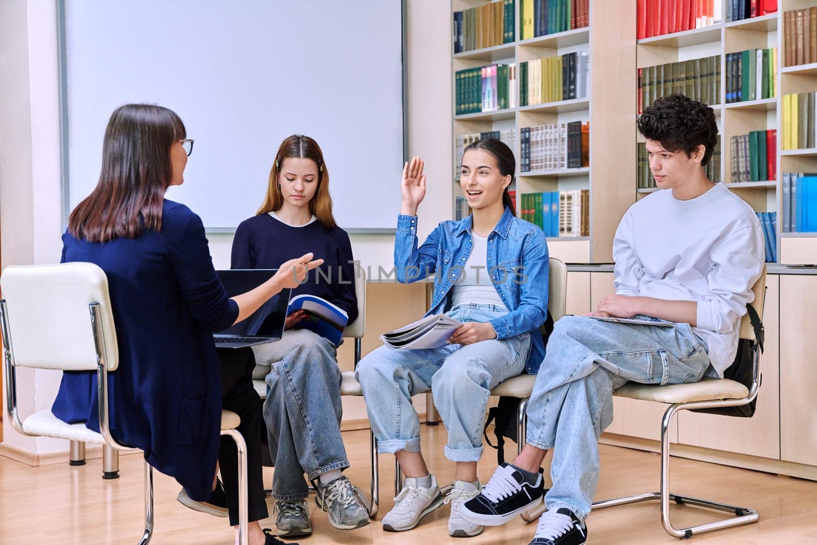 Group of teenage students sitting with female mentor teacher psychologist counselor social worker inside classroom library office. High school education teamwork learning behavior adolescence concept