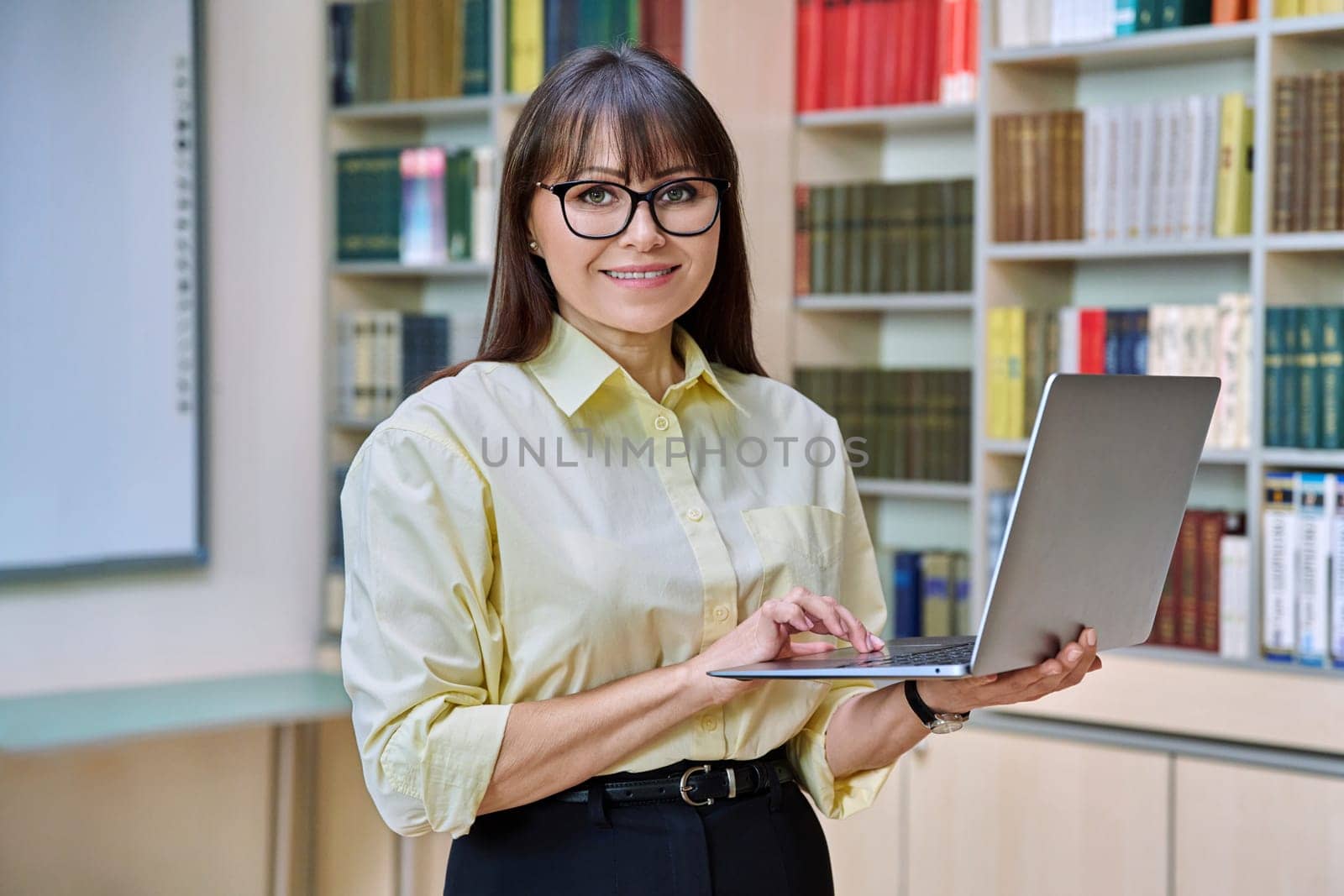 Middle aged elegant woman using laptop computer inside library. Confident smiling elegant business female looking at camera teacher mentor professor leader manager psychologist counselor social worker