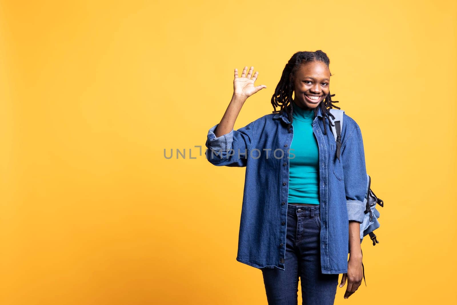 Joyful african american woman greeting school colleagues, isolated over yellow studio background. Happy student holding backpack waving hand, saluting mates, showing positive mood