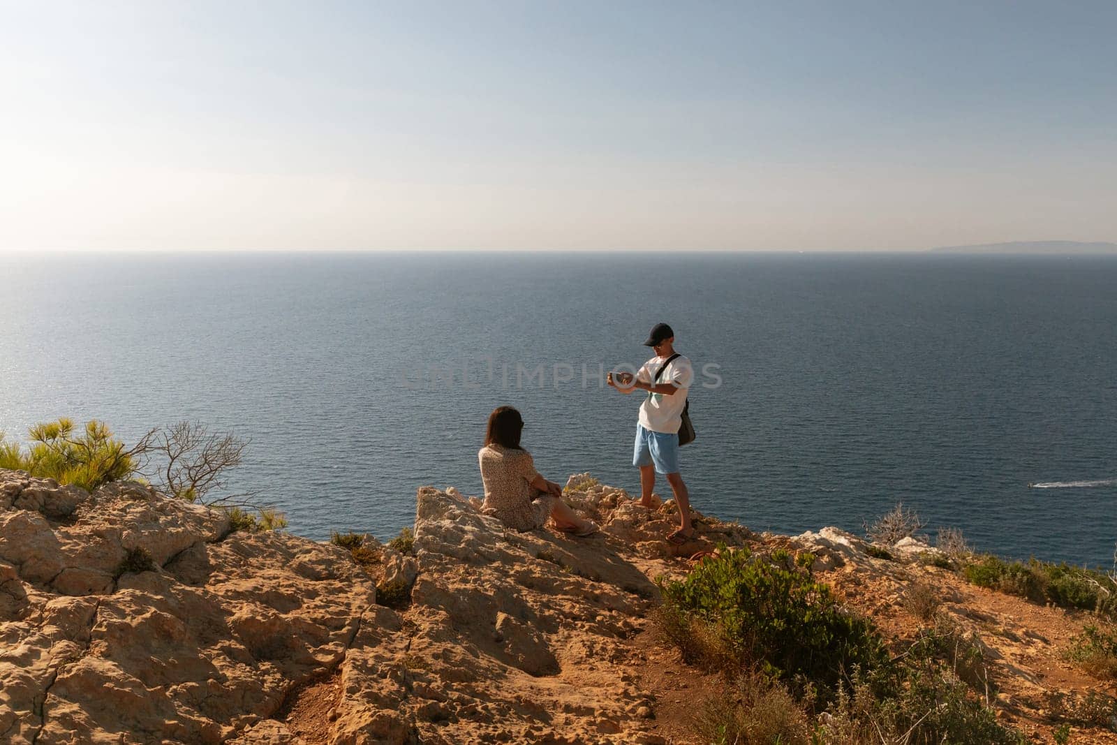 One cool guy in a cap takes a smartphone photo of a girl in a dress sitting on the top of a mountain against the backdrop of a blurred sea on a sunny summer day, close-up side view.