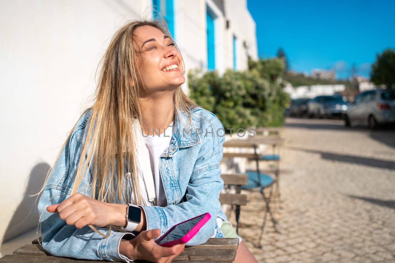 Beautiful blond woman holding smartphone at outdoor cafe table. Happy female tourist in denim shirt sitting near white building. Girl enjoys her vacation in typical Portugal city.
