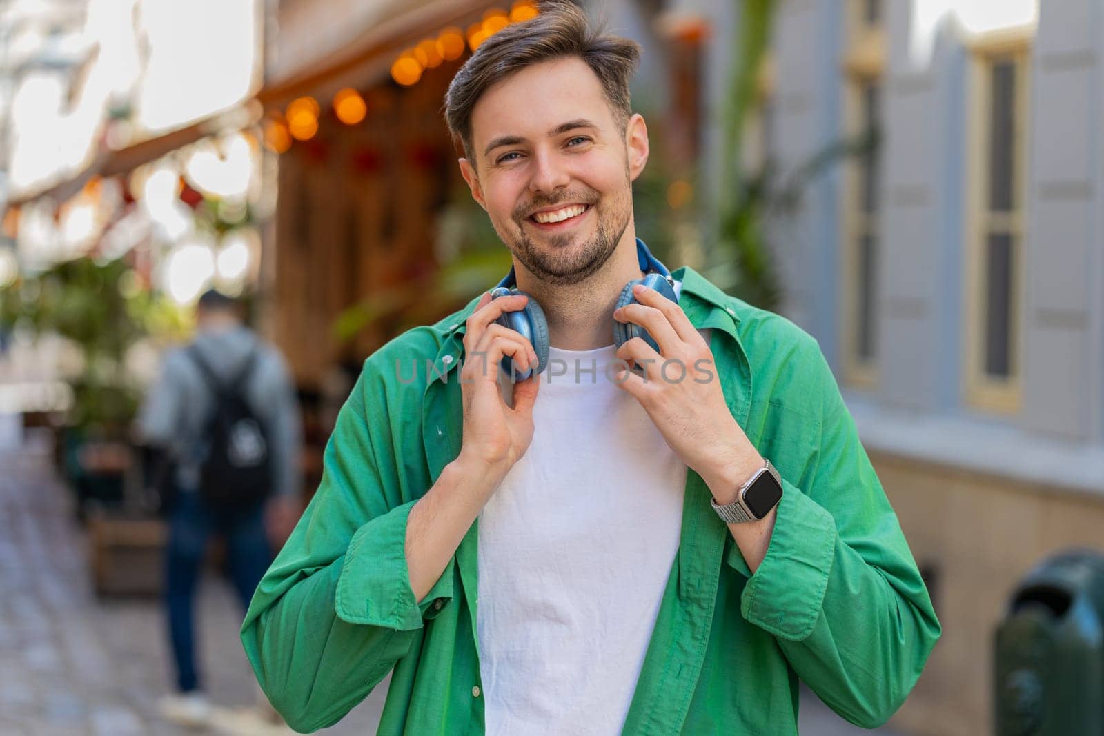Portrait of smiling young male tourist in casual clothes with wireless headphones around neck. Happy positive Caucasian man traveler standing on city urban street looking at camera. Town lifestyles.
