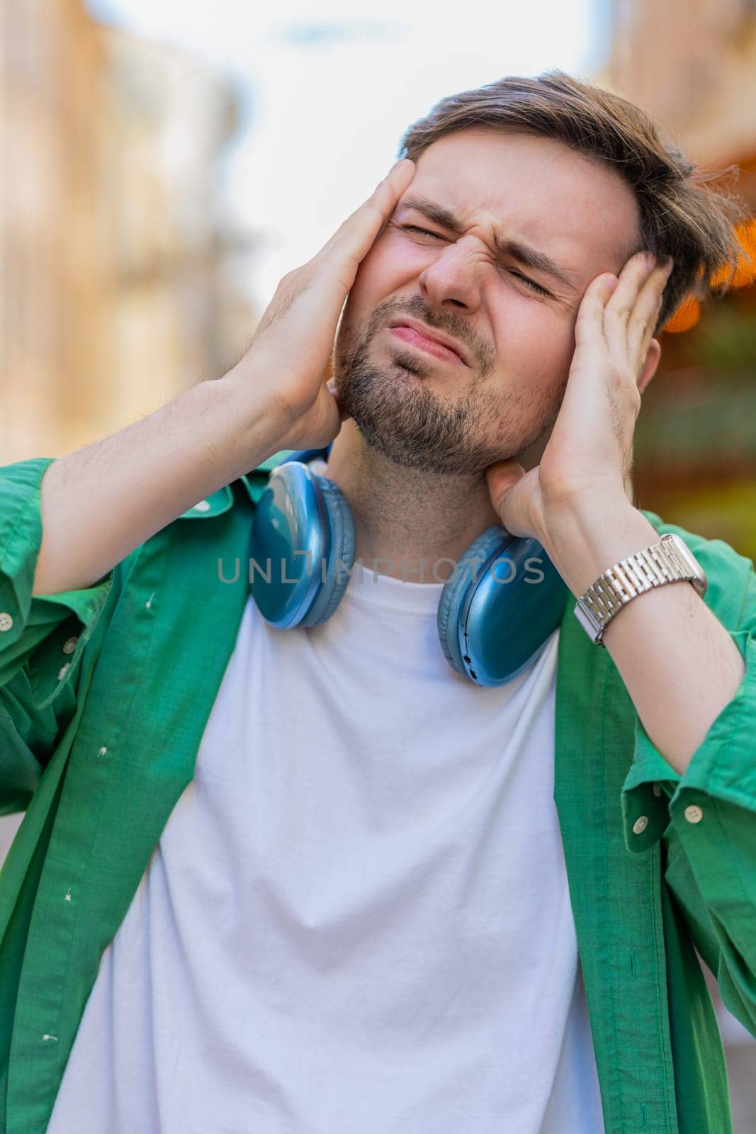 Displeased exhausted Caucasian man rubbing temples to cure headache problem suffering from tension migraine stress. Young guy tourist grimacing, pain high blood pressure standing on city urban street.