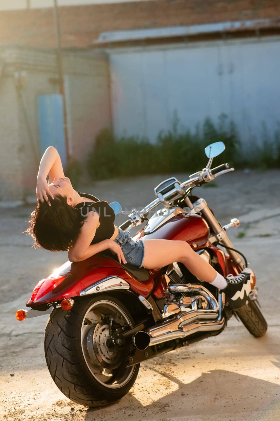 A brunette in shorts poses while lying on a red motorcycle. Vertical photo