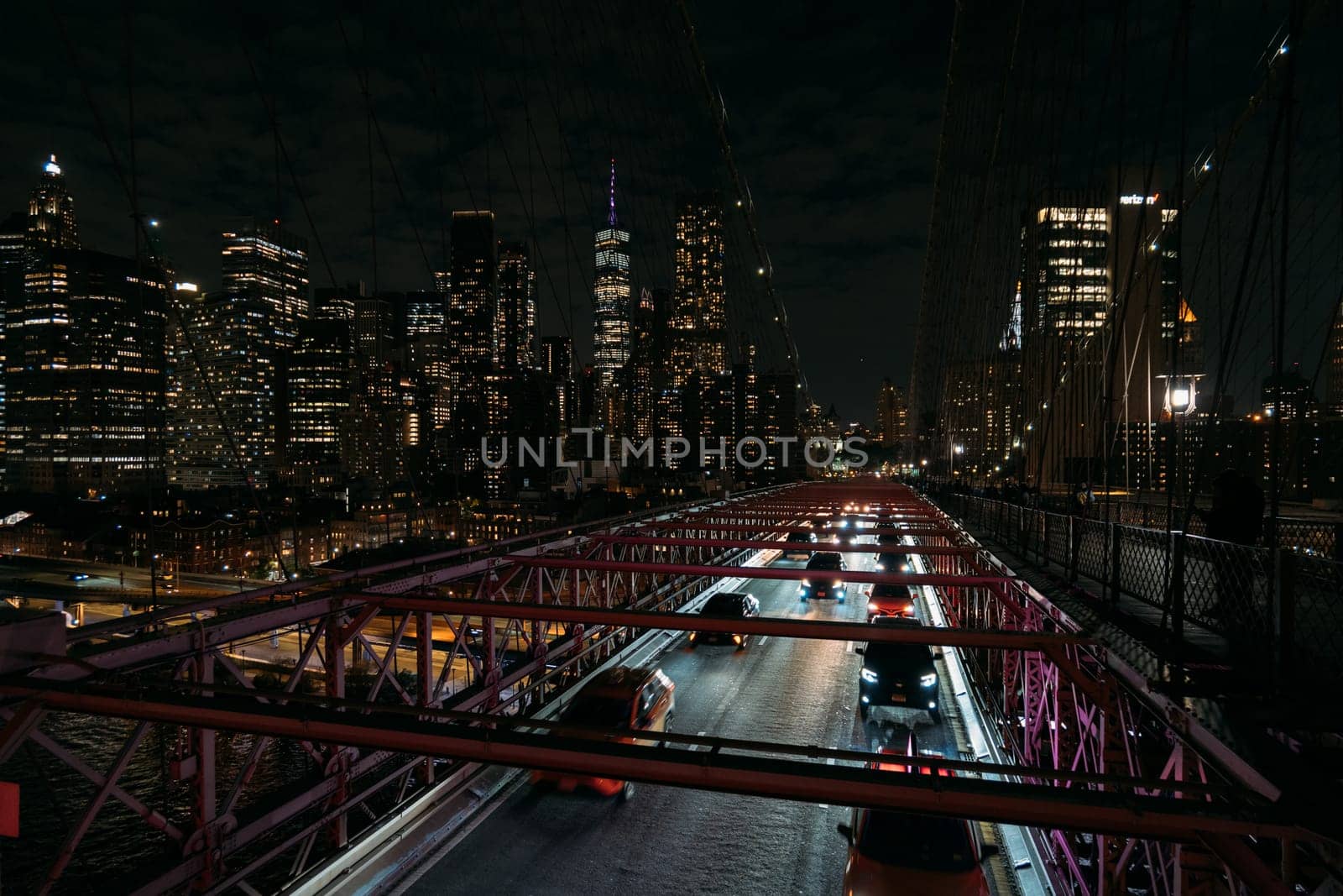 View of cars moving on the Brooklyn Bridge at night with the illuminated skyline of New York City in the background. The photograph captures the busy city life and iconic landmarks.