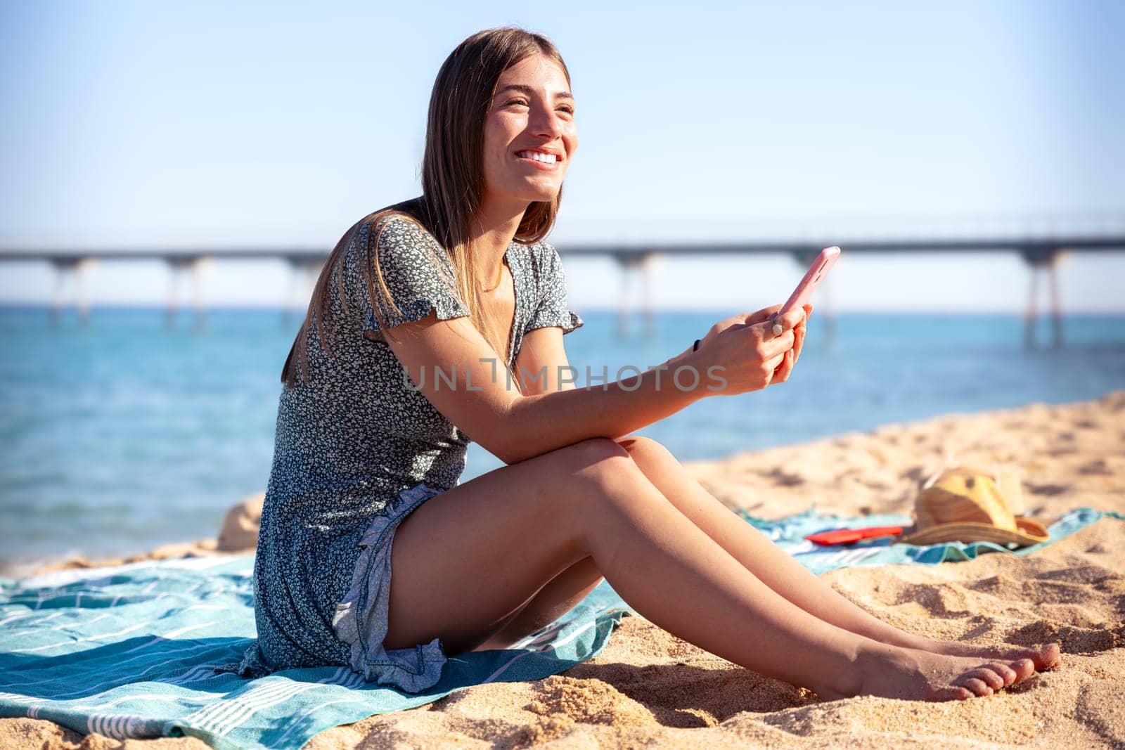 young happy caucasian woman on vacation using smartphone app sitting on towel on the beach