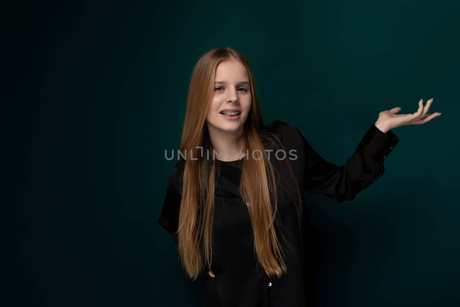 A woman with vibrant, long red hair stands confidently in front of a solid green wall, her presence commanding attention. Her hair cascades down her back, drawing eyes towards her elegant posture.