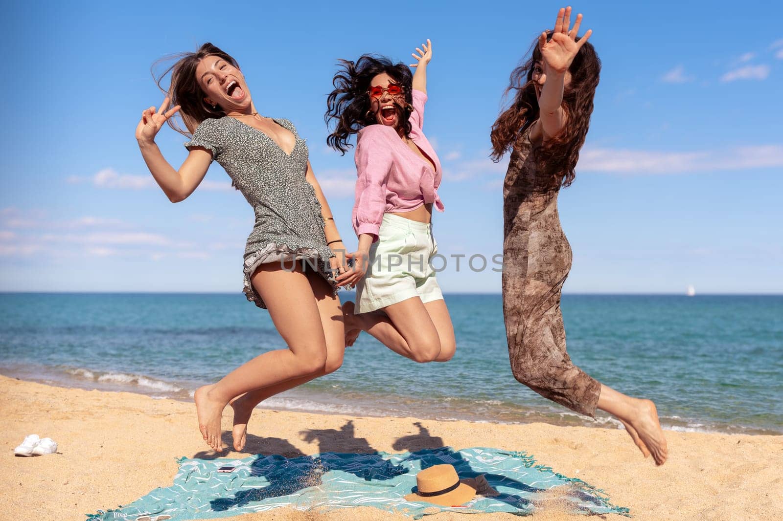 Three happy girls in jumping motion with on the beach outdoors in summer celebrating vacation days. Friends having fun on vacation by the sea carefree, and party concept