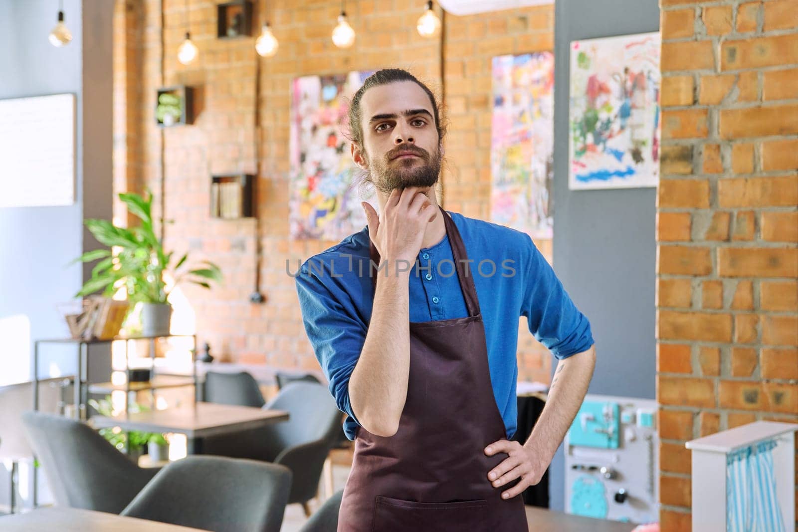 Confident successful young man service worker owner in apron posing looking at camera in restaurant cafeteria coffee pastry shop interior. Small business staff occupation entrepreneur work