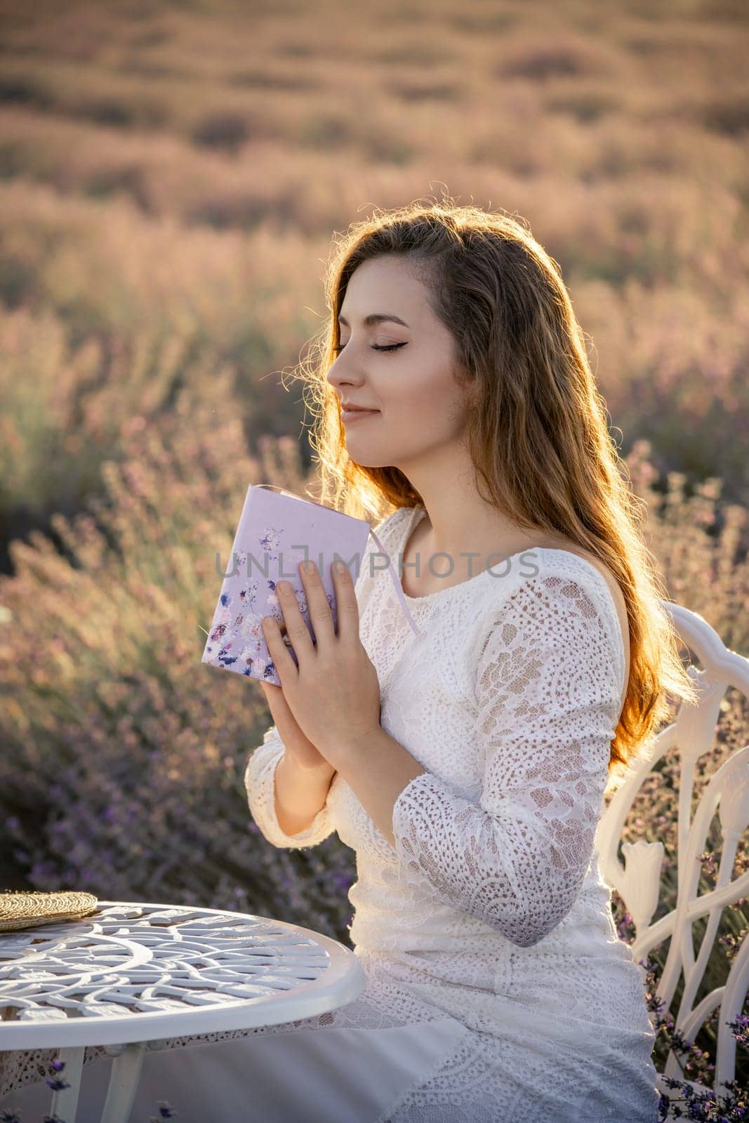 A woman in a white dress is sitting at a table with a book in her lap. She is in a peaceful and relaxed state, possibly reading the book or meditating. The scene is set in a field of lavender