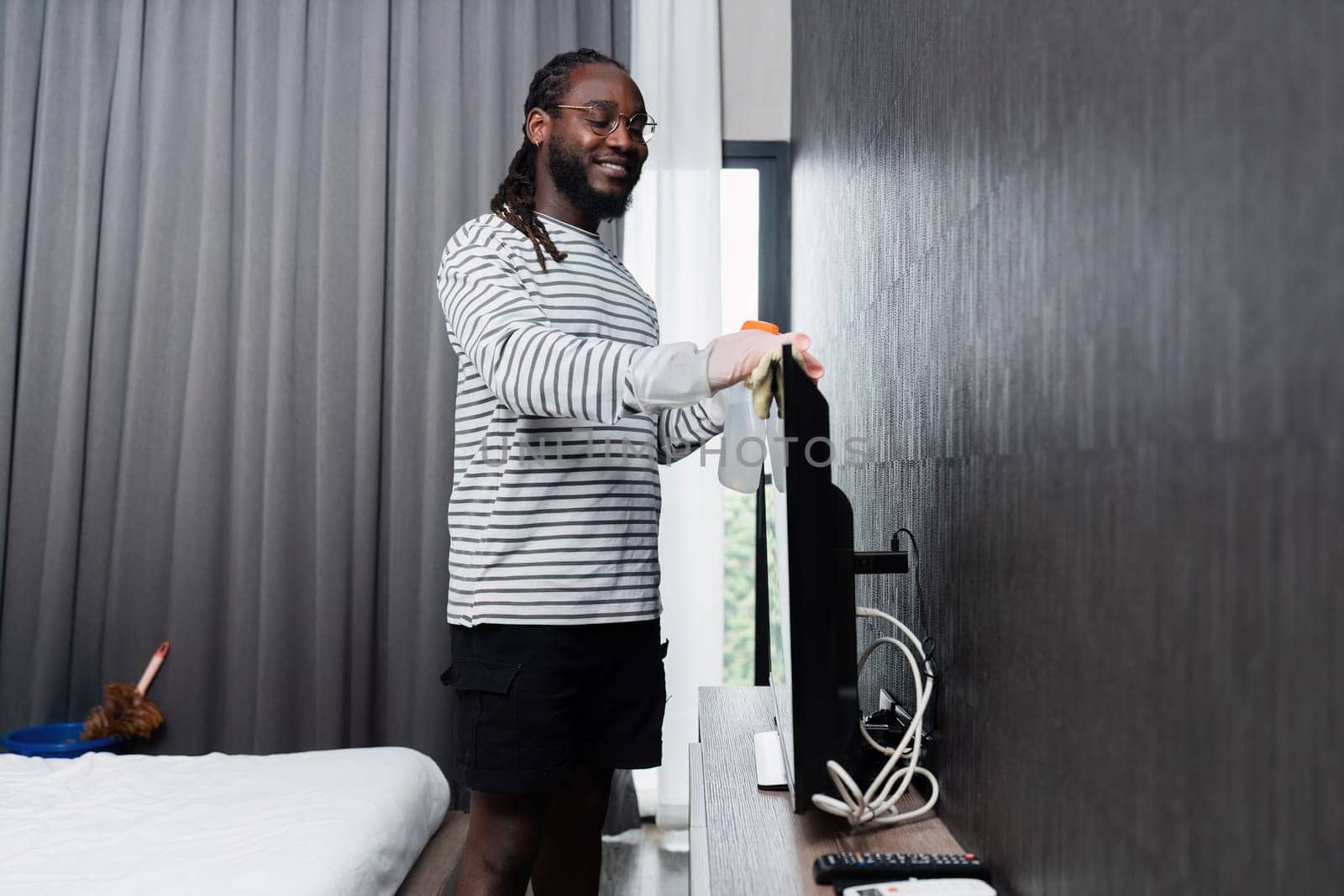 Man dusting TV screen in a modern bedroom, ensuring a clean and tidy living space. Home cleaning and maintenance concept.