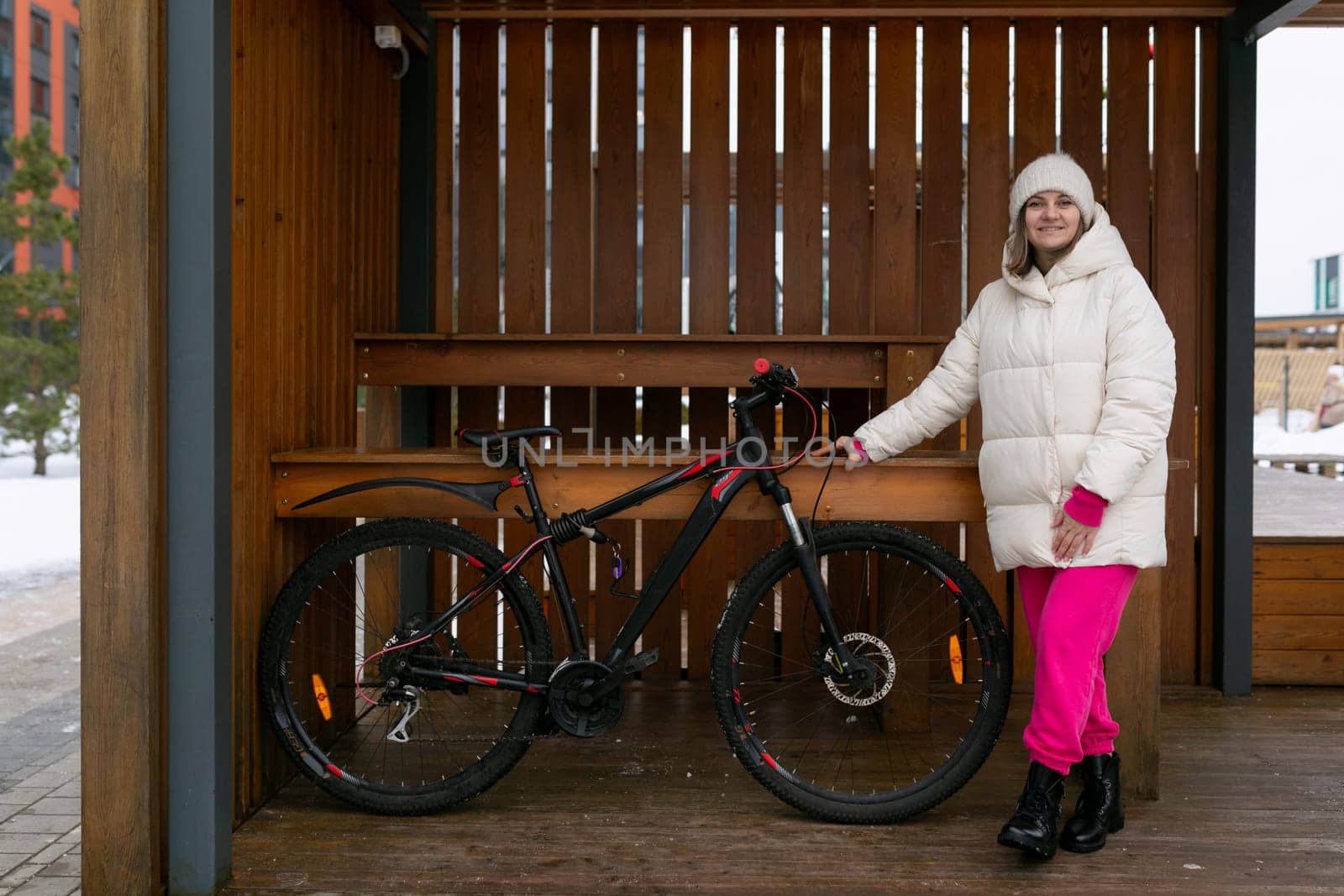 A woman is standing next to a bicycle in a sheltered area, possibly a bike storage or parking facility. She appears to be adjusting something on the bike or its accessories. The setting seems to be a functional space for bike riders to safely store or work on their bicycles.