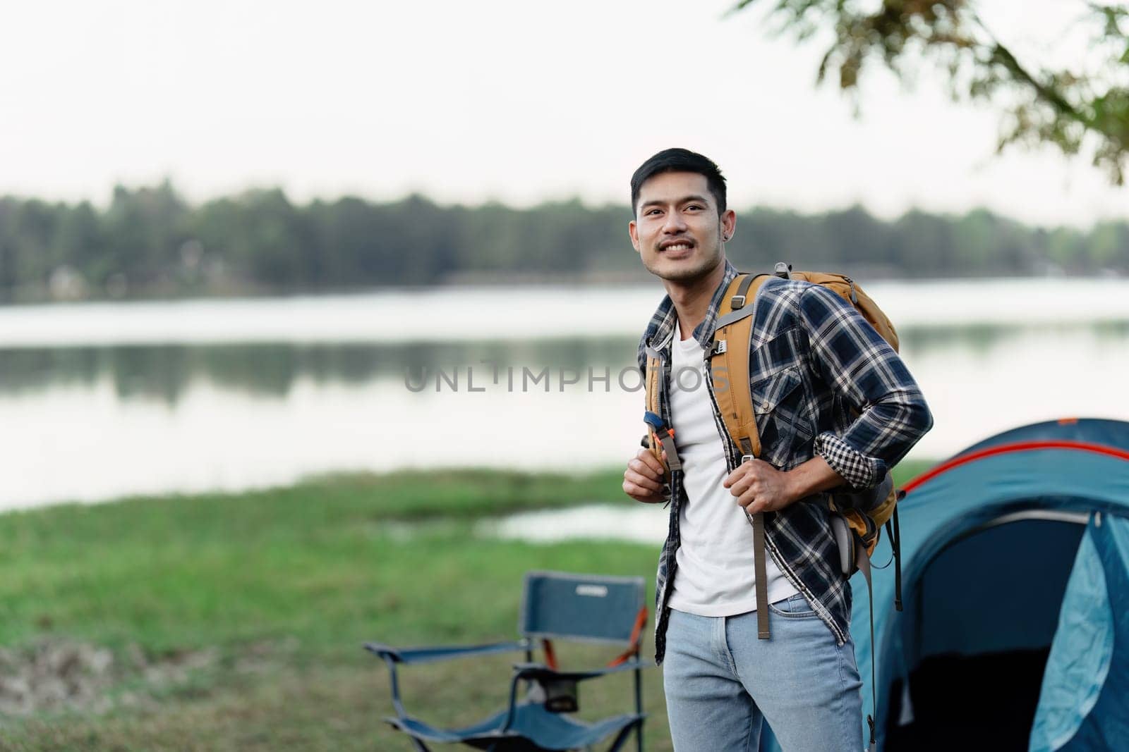 A young man with a backpack stands near a tent by the lake, ready for a travel camping adventure in a beautiful outdoor setting.