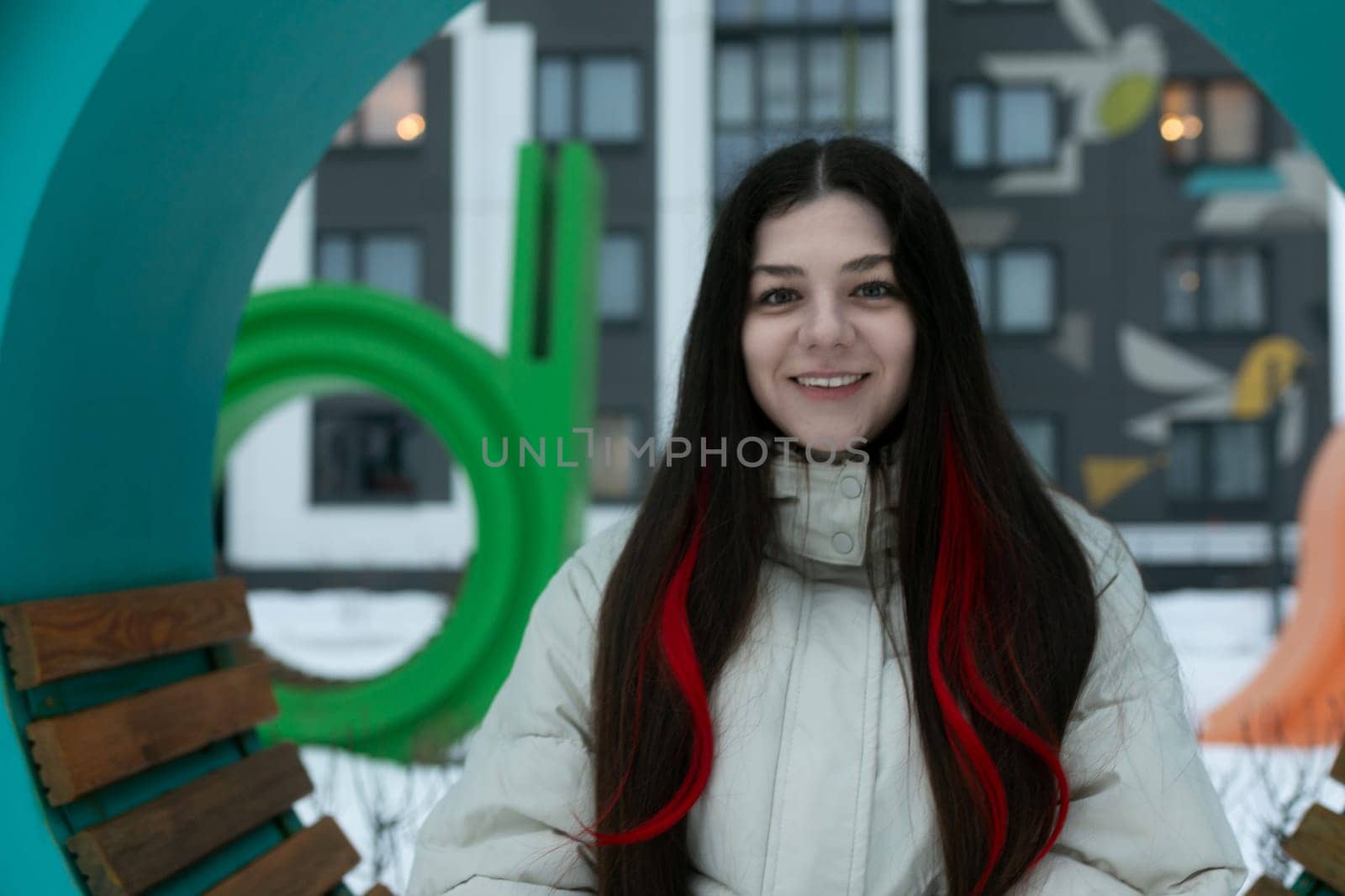 A woman with long hair is standing in front of a wooden bench, looking towards the camera. She is wearing casual clothing and appears relaxed in the outdoor setting.